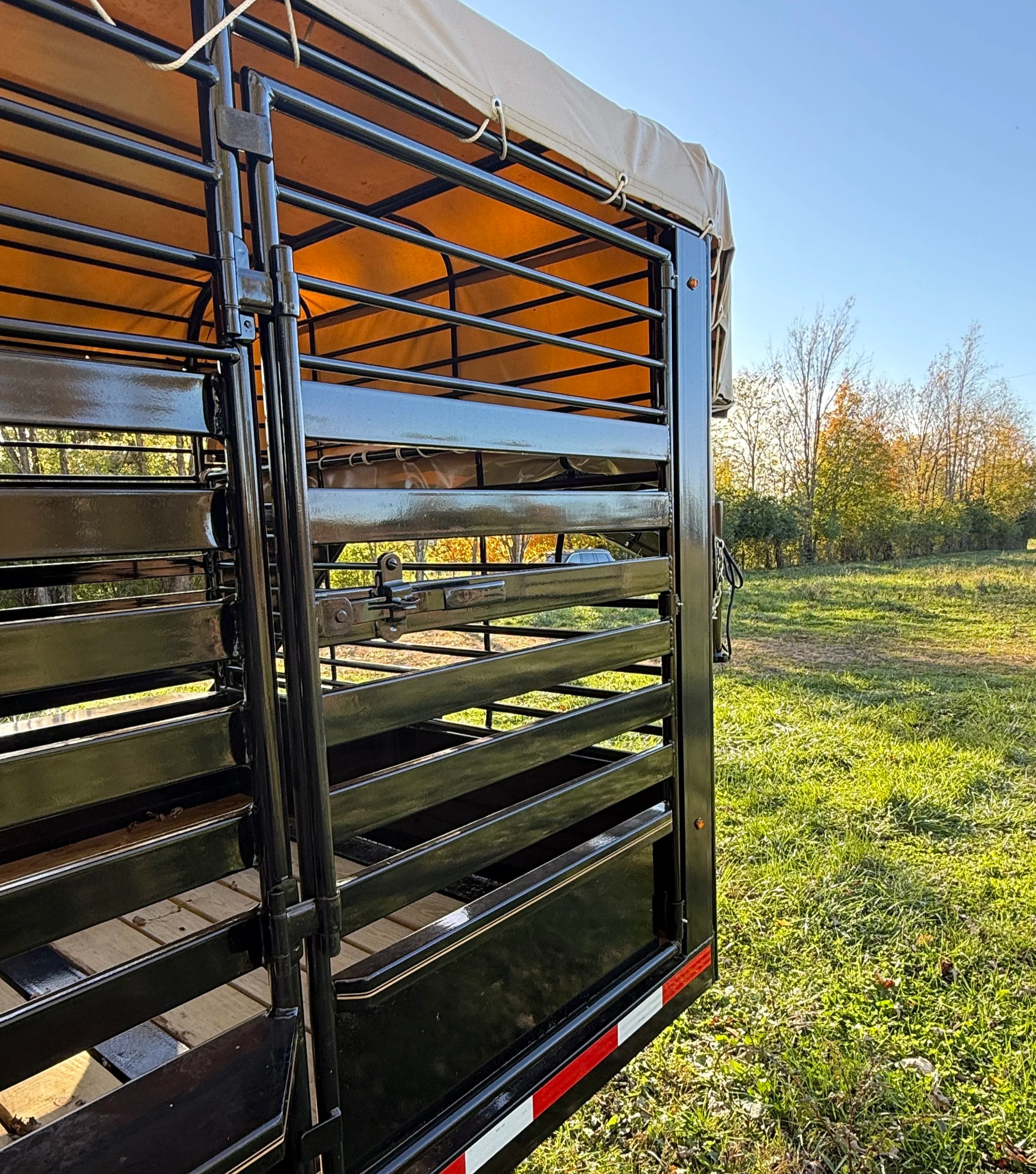 Black utility trailer with open sides and a tan canopy, standing on grass with trees and a blue sky in the background.