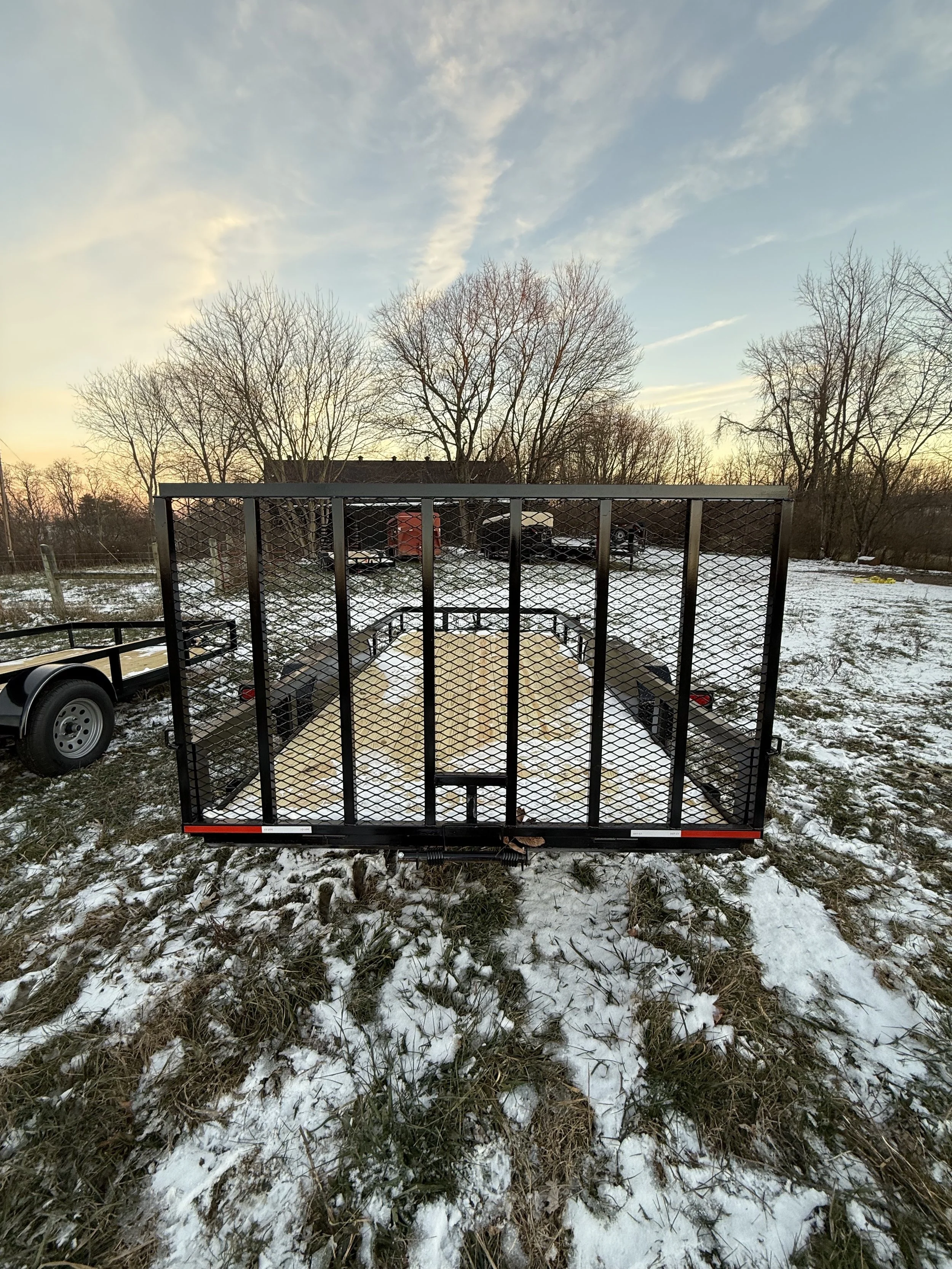 A black utility trailer with a metal mesh ramp gate, parked on a snowy grassy field during sunset, with leafless trees and a cloudy sky in the background.