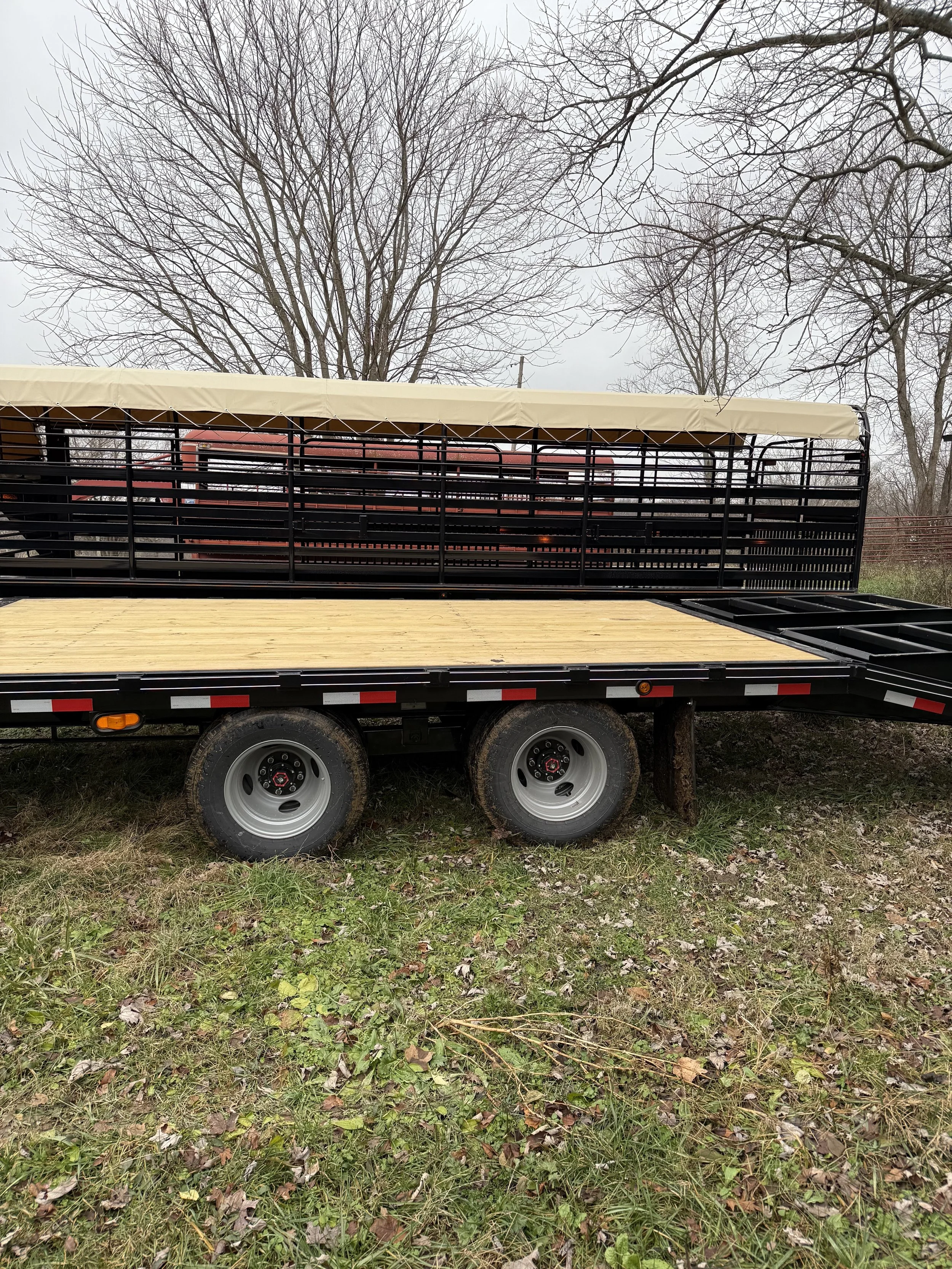A flatbed trailer with two wheels parked on grass, with a black metal side rail and a beige canopy, and leafless trees in the background.