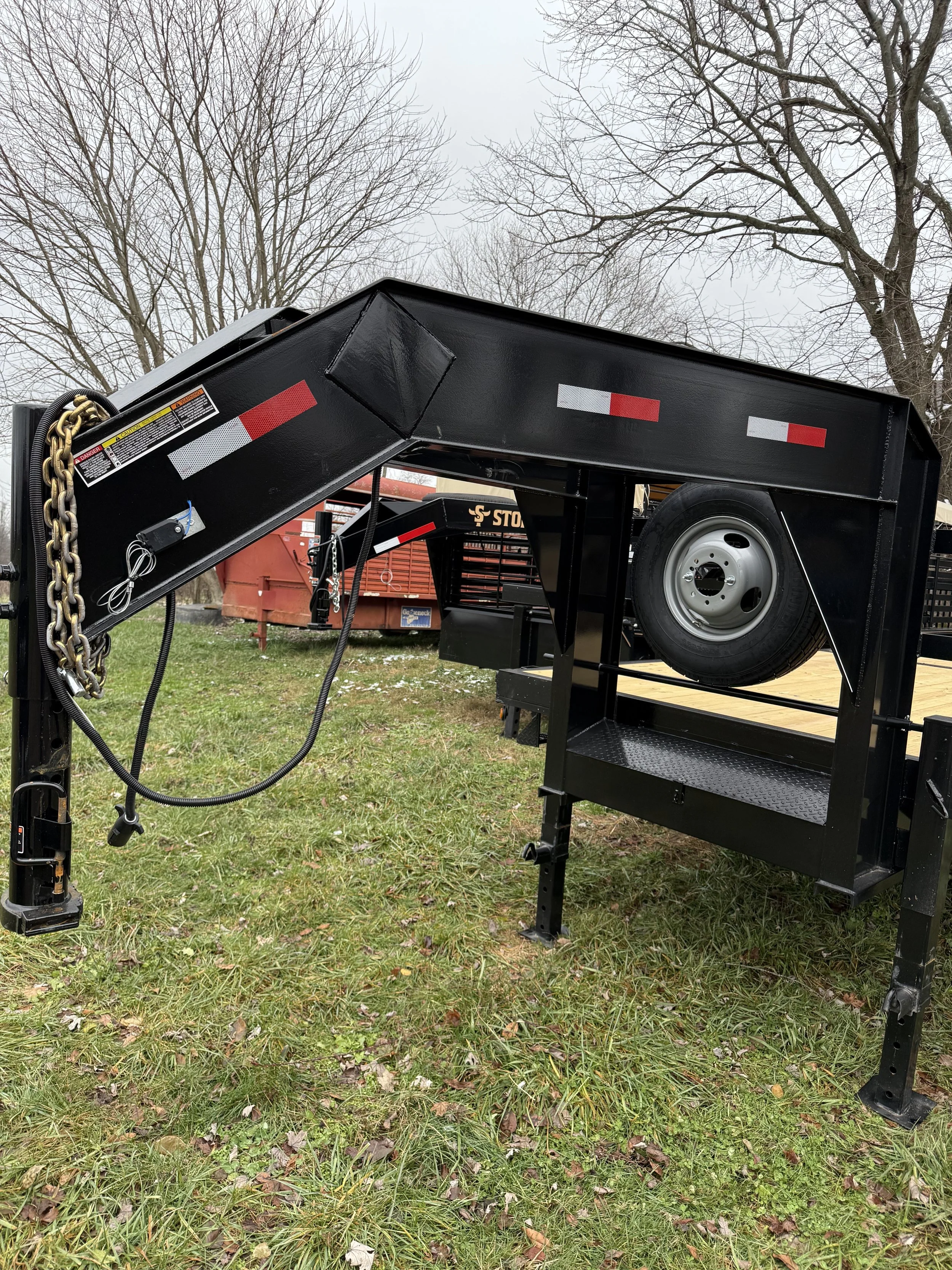 Black flatbed trailer with a spare tire mounted on its side, parked on grass with trees in the background.