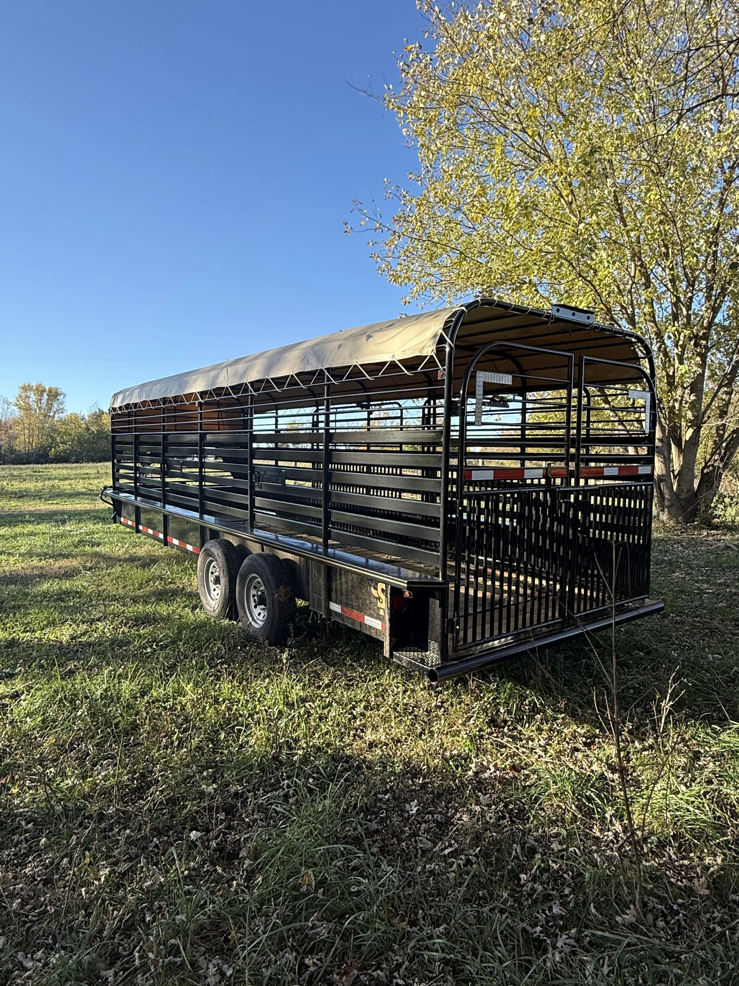 Black livestock trailer with a beige canopy, parked on grassy land with trees and a clear blue sky in the background.