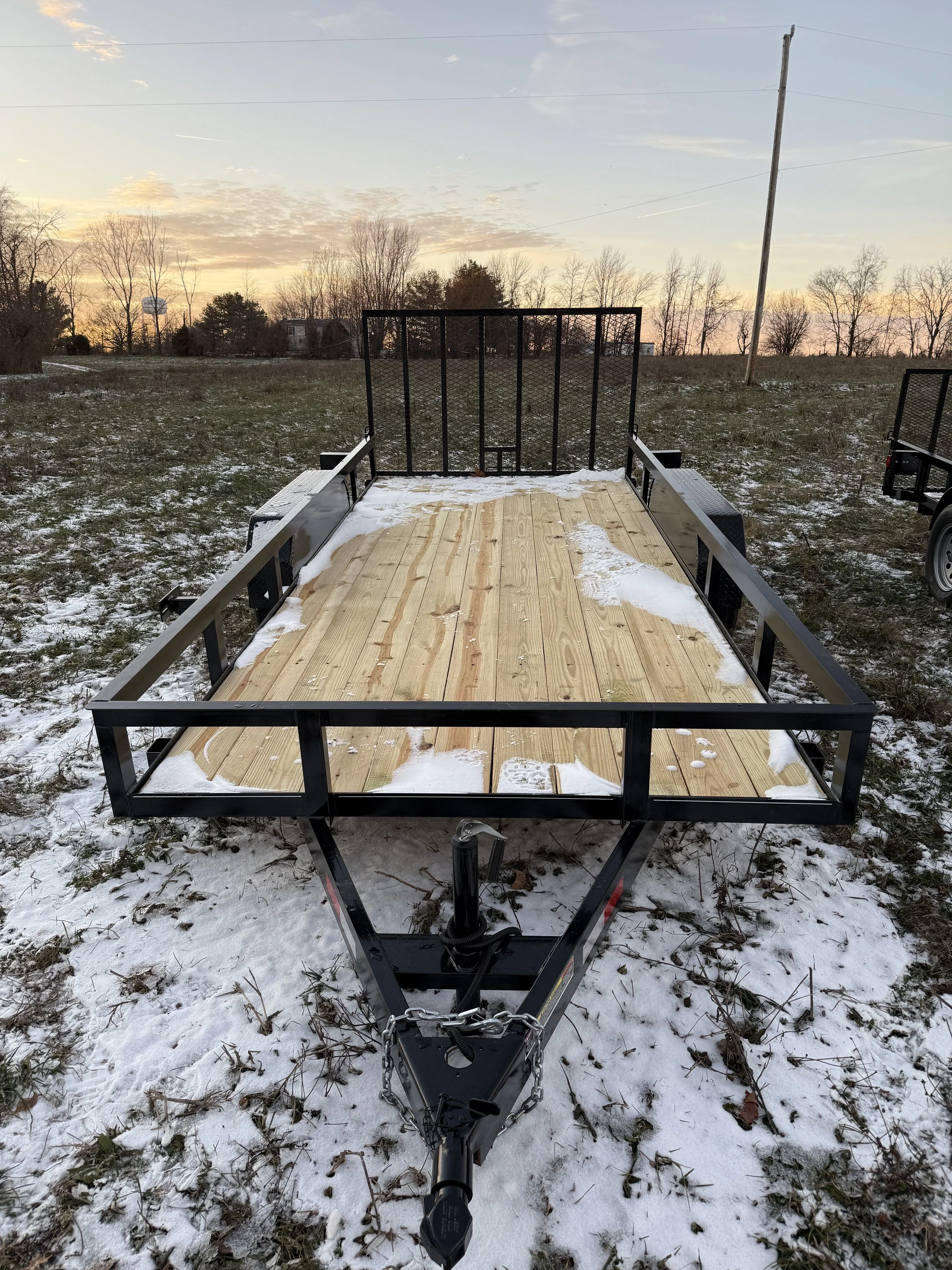 A flatbed utility trailer with a wooden deck, black metal frame, and safety rails, parked on a snowy field during sunset.
