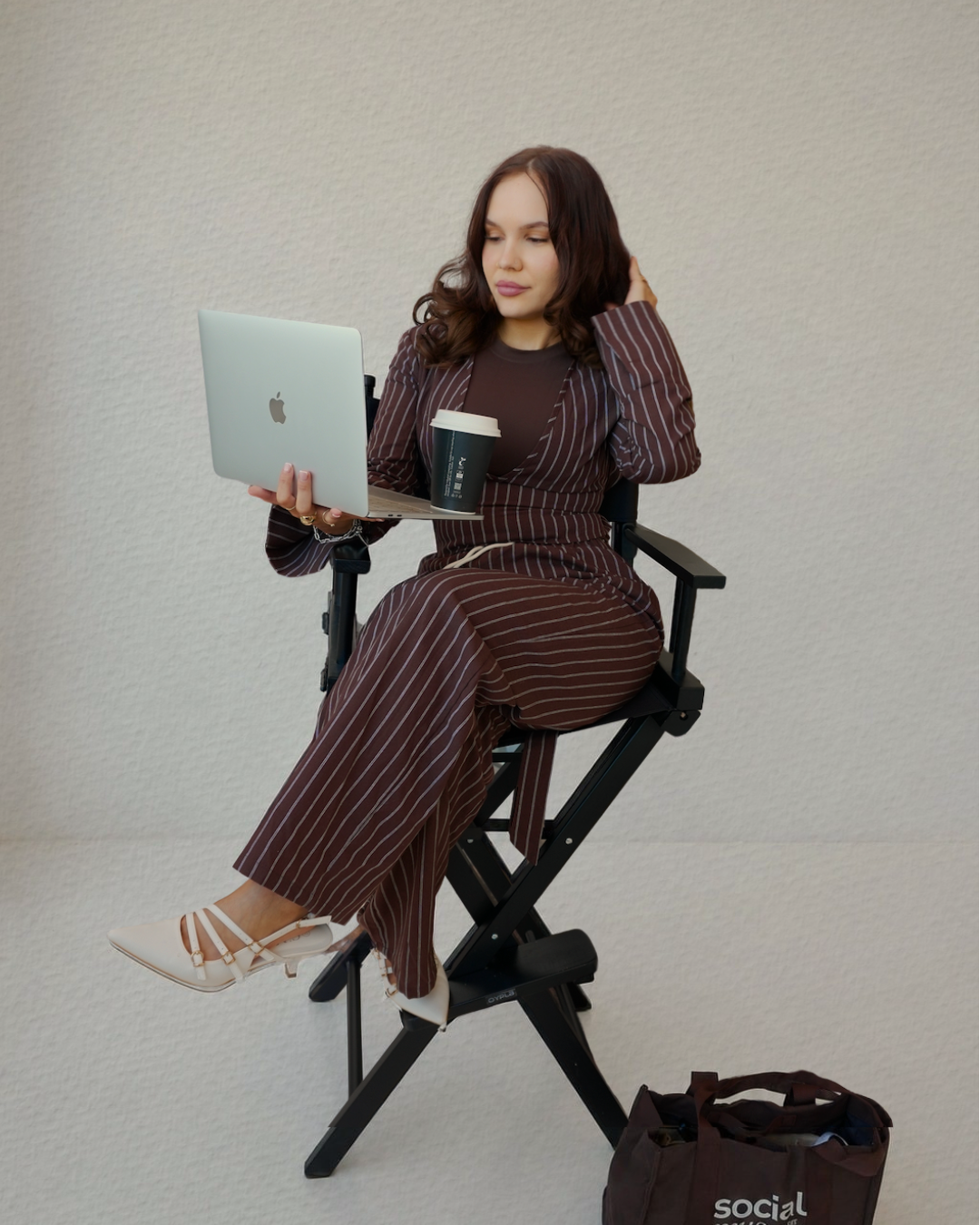Woman in striped pants and blazer sitting on a director's chair, working on a laptop, with a coffee cup in her lap and a bag on the ground.