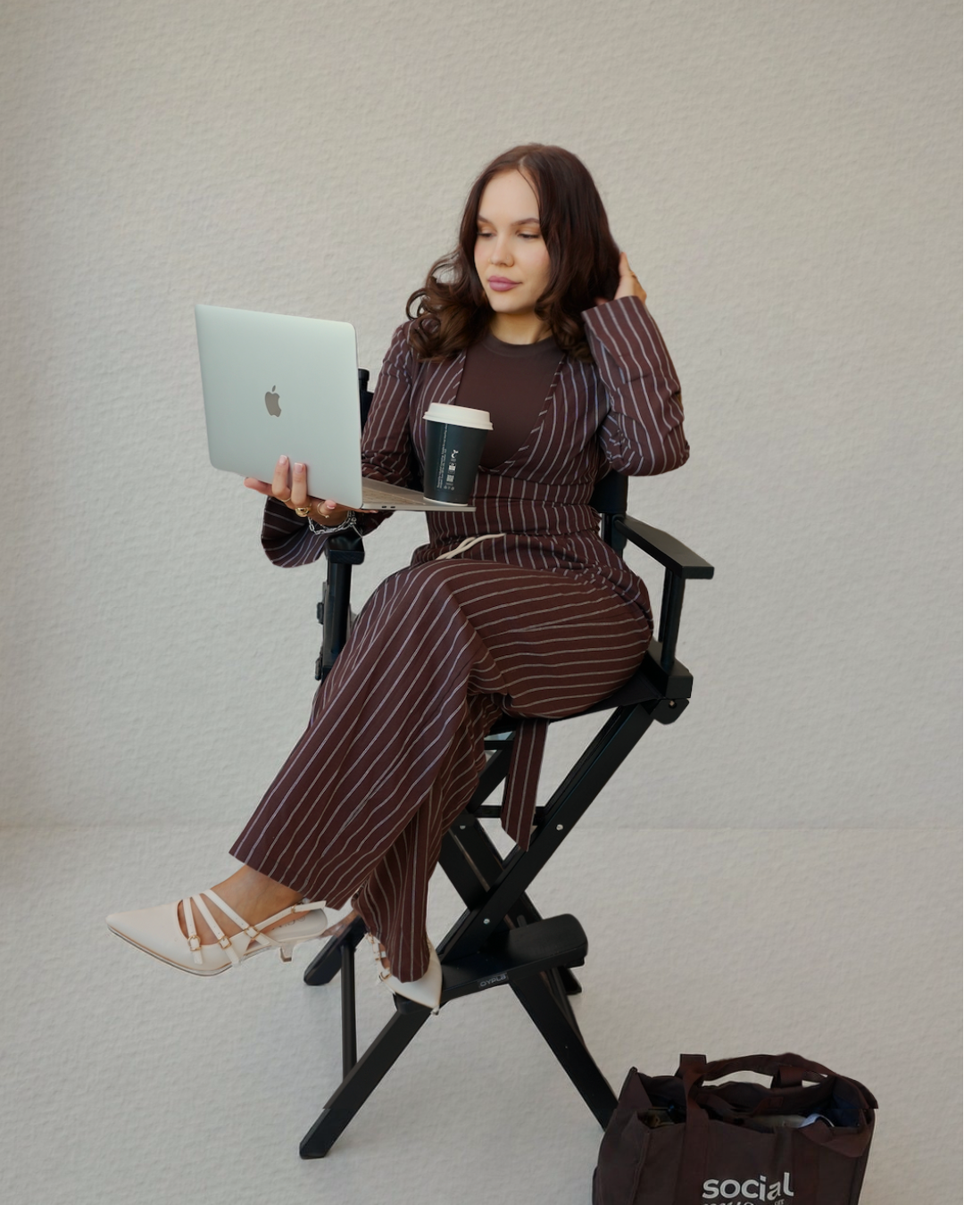 Woman in pinstripe suit sitting on director's chair, holding a laptop and a coffee cup, with a bag on the floor nearby.