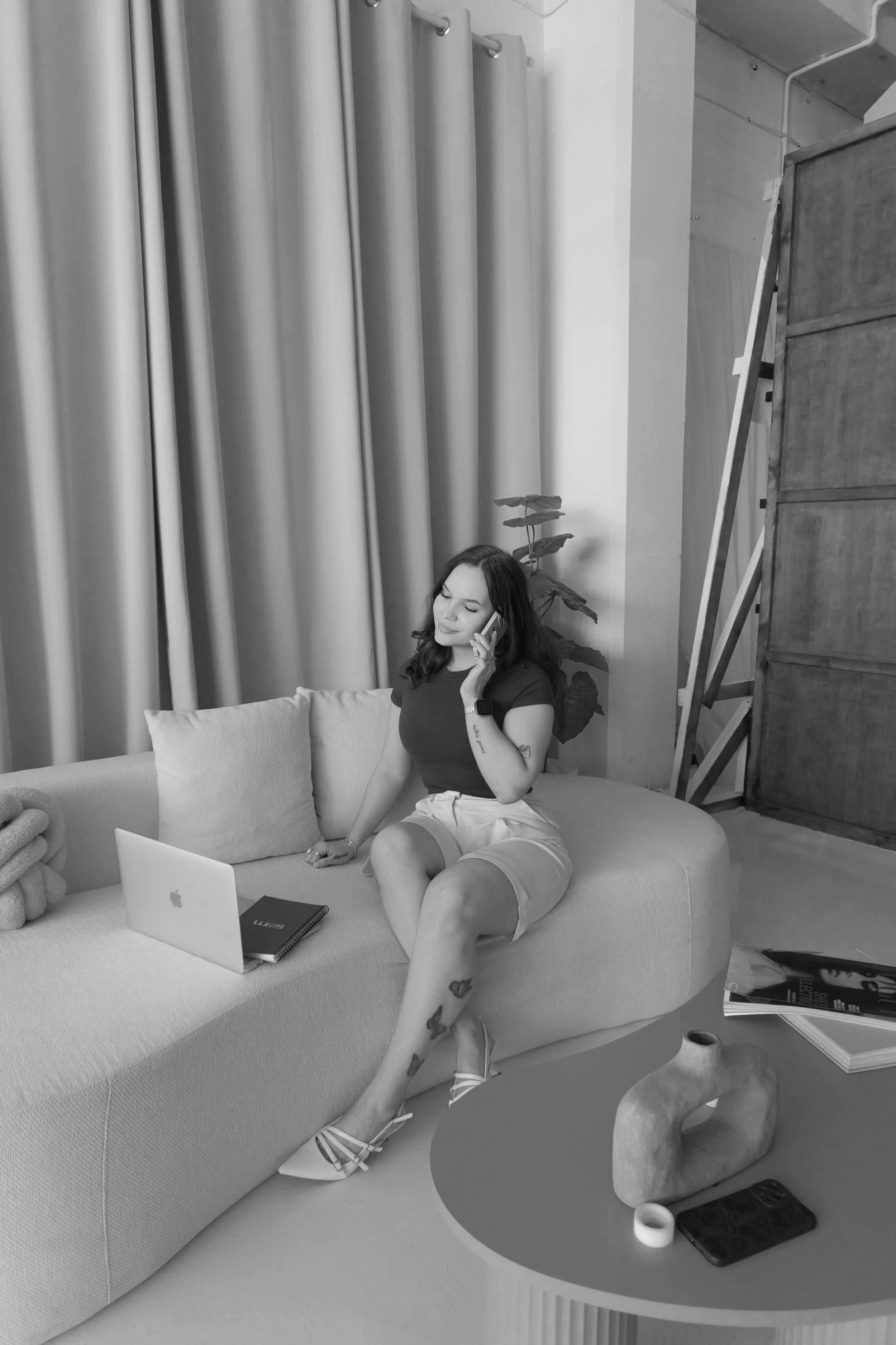 A woman sitting on a couch in a modern living space, talking on her mobile phone. There is a laptop, a notebook, and a pen on the couch beside her. She is wearing a dark top and light shorts, with heels. Behind her, a window with curtains, a potted plant, and a wooden cabinet are visible. A small round table near her holds candles, a remote control, and a rolled-up magazine.