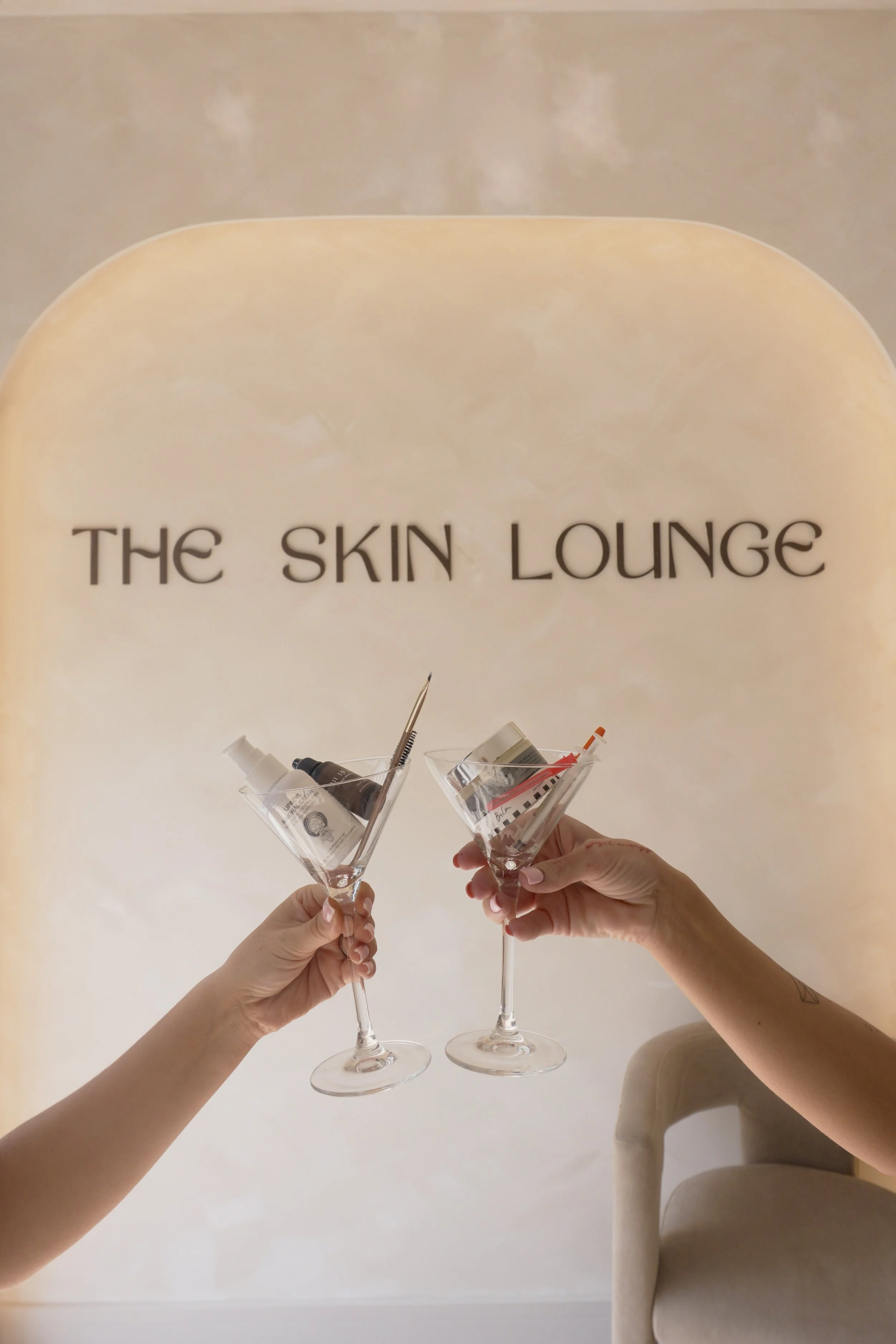 Two hands holding champagne glasses with skincare products inside, raised in a toast in front of a sign that reads 'The Skin Lounge' on a beige wall.