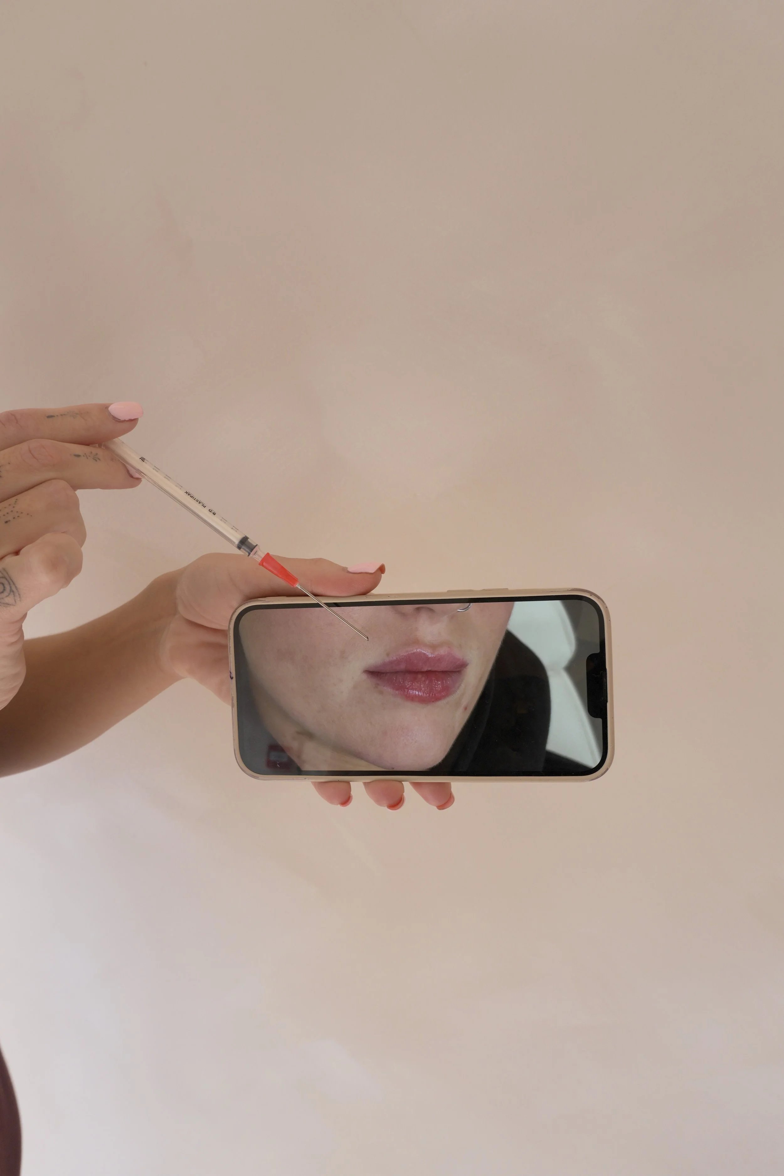 Person holding a smartphone with a mirror reflection of their face, as they prepare to give themselves an injection with a syringe, against a plain light-colored background.