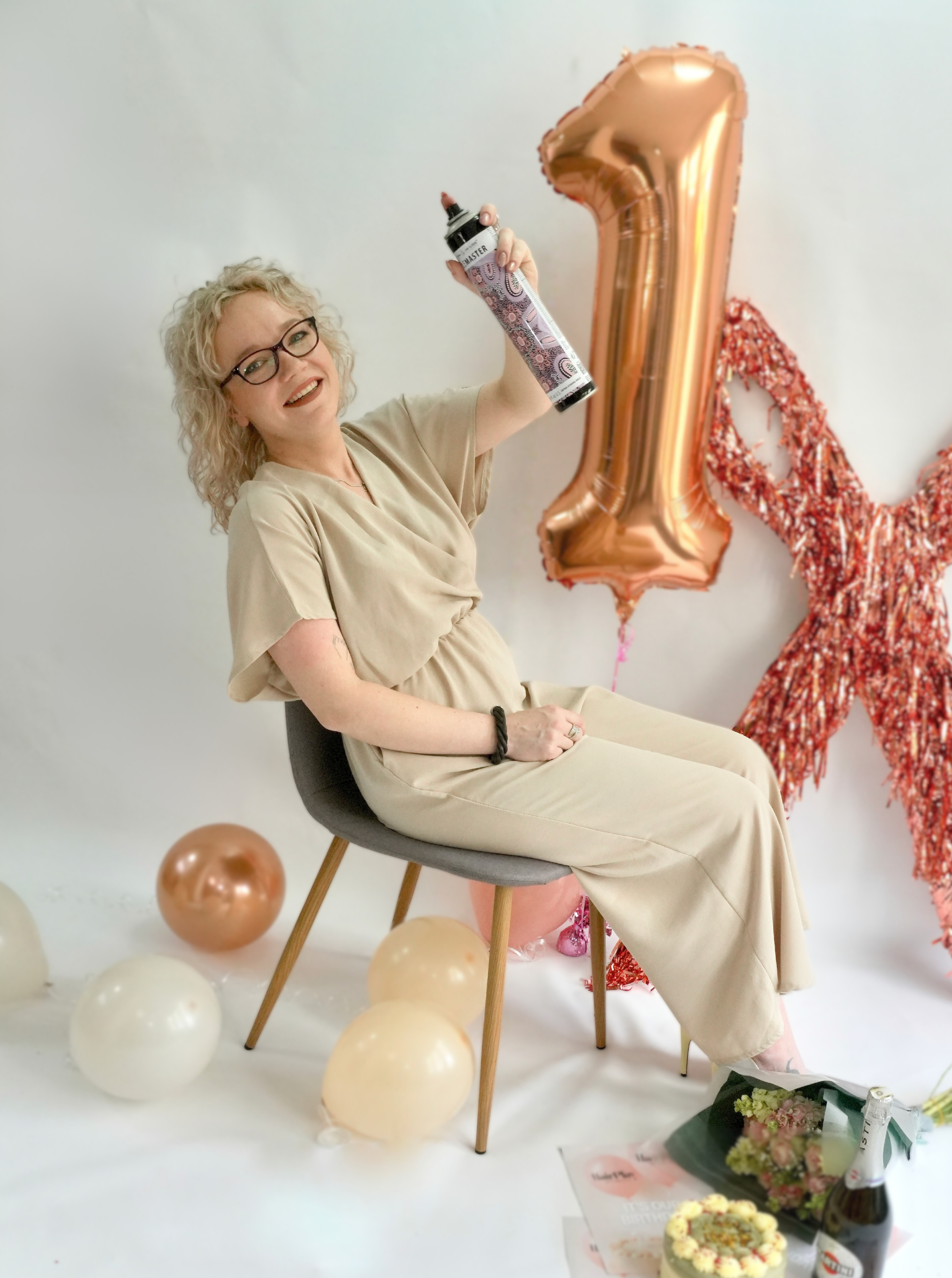 A woman with curly blonde hair and glasses sitting on a chair, holding a black marker with a decorative cover, surrounded by white, cream, and rose gold balloons. Behind her is a large rose gold number one balloon and a red garland. On the floor in front of her are flowers, a cake, and bottles of champagne.