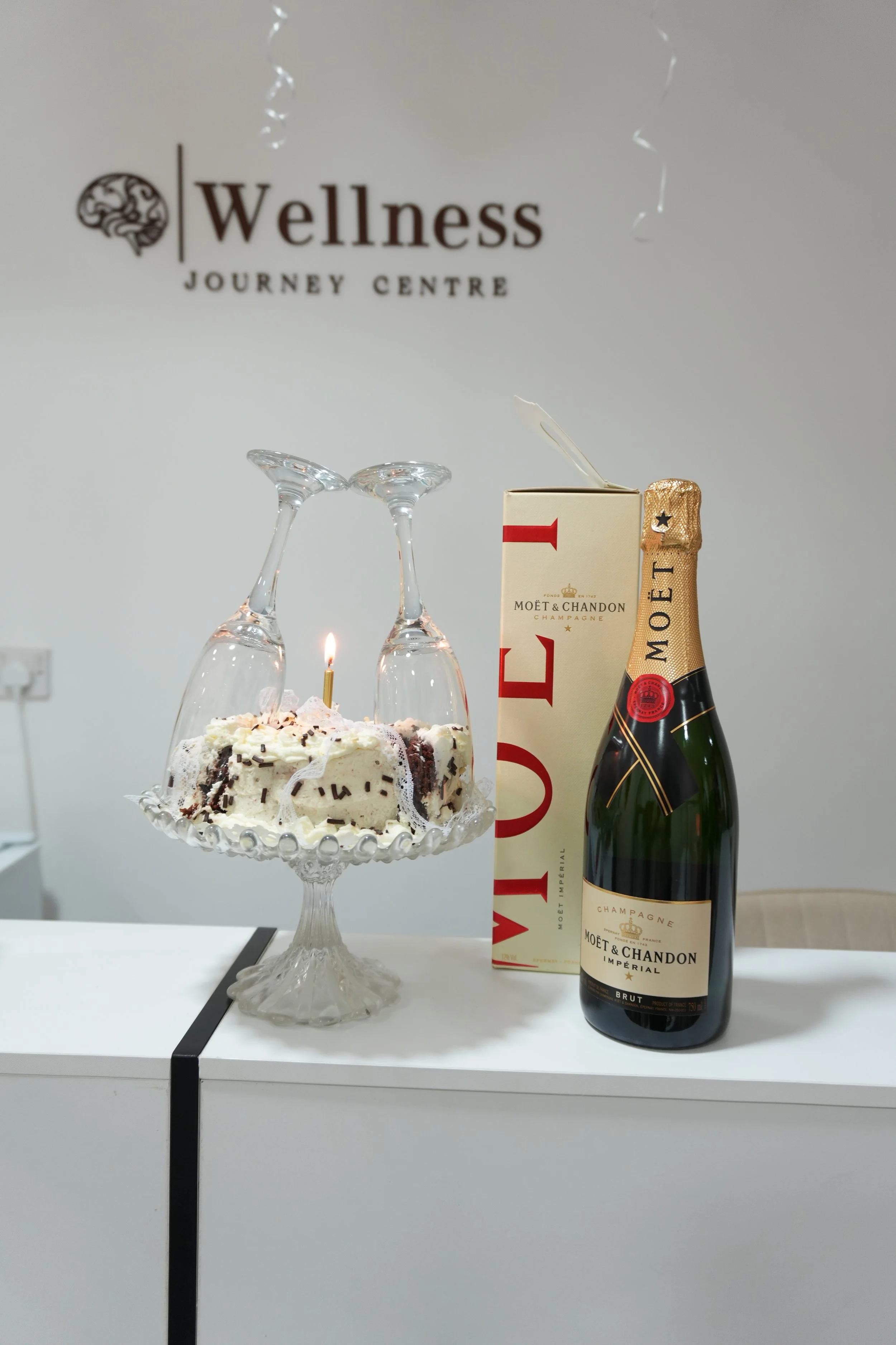 A cake with a lit candle and two upside-down wine glasses on a glass cake stand, surrounded by a bottle of Moët & Chandon champagne and its packaging, with a sign in the background reading 'Wellness Journey Centre'.