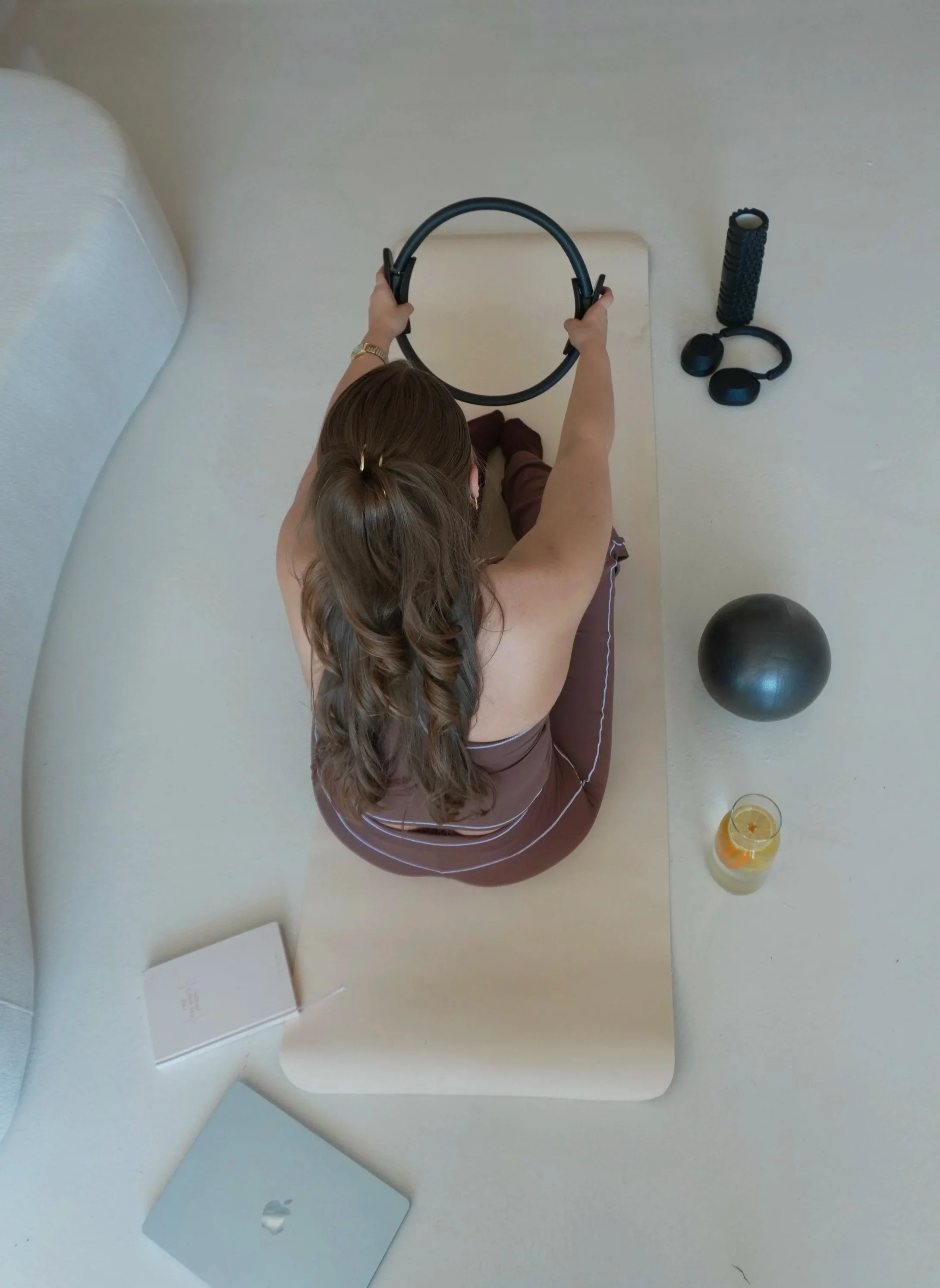 Top-down view of a woman holding a circular fitness ring, with a laptop, a glass of lemonade with ice and fruit, fitness equipment including a foam roller, a massage ball, and a pair of headphones on a white desk or table.