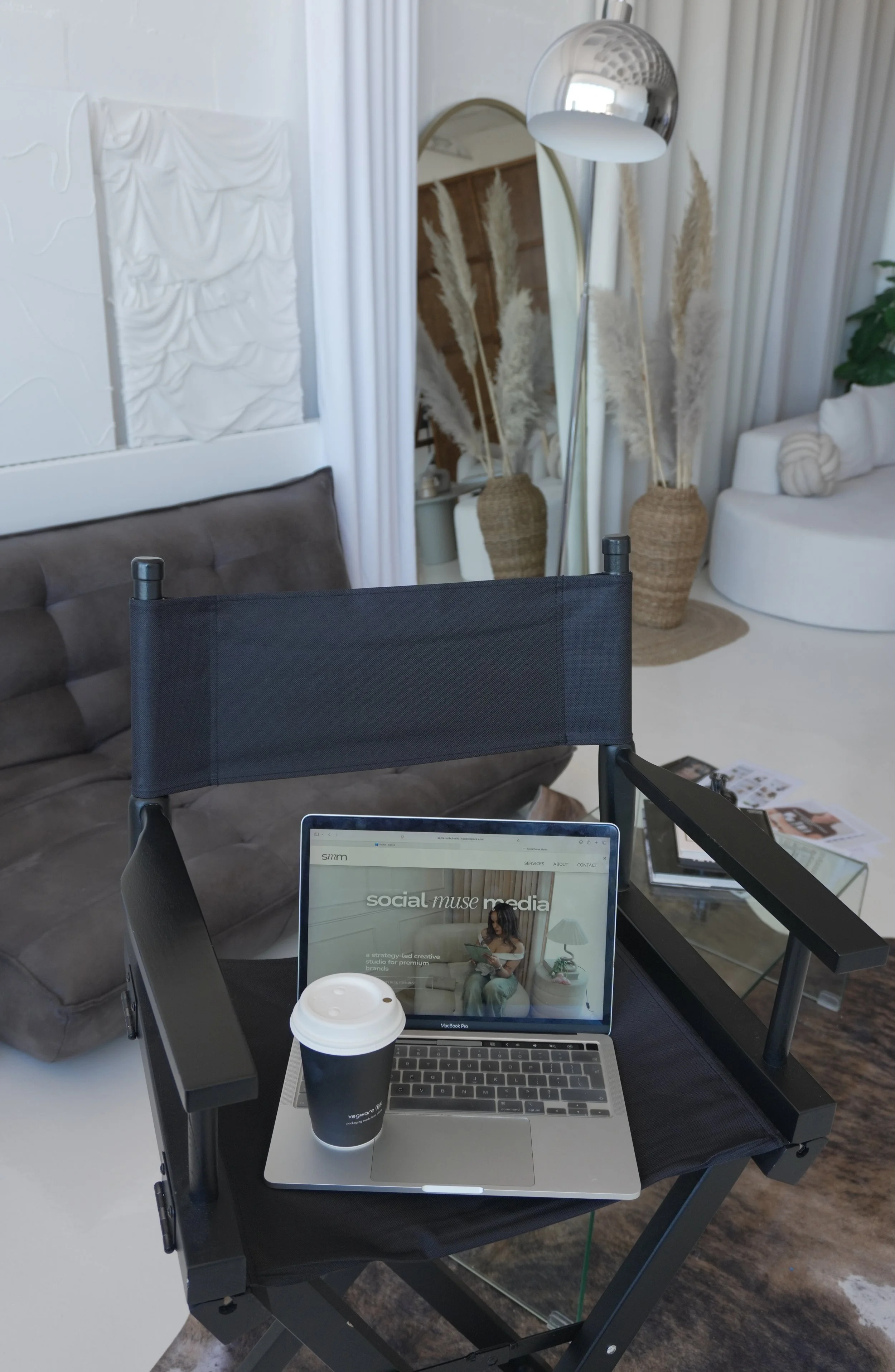 A workspace with a laptop displaying a website for social media strategy, a paper coffee cup, and a black coffee cup on a table. The background shows a mirror, plants in baskets, white curtains, a gray couch, and a floor lamp.