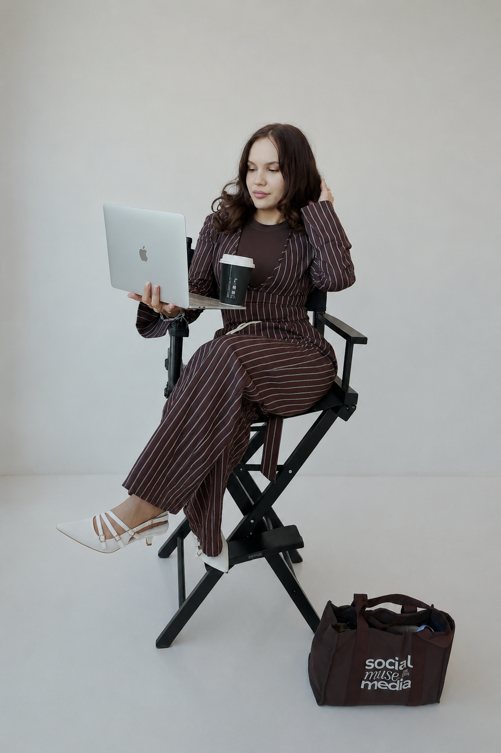 A woman sitting on a director's chair, looking at her laptop, holding a coffee cup, with a brown bag on the floor nearby.