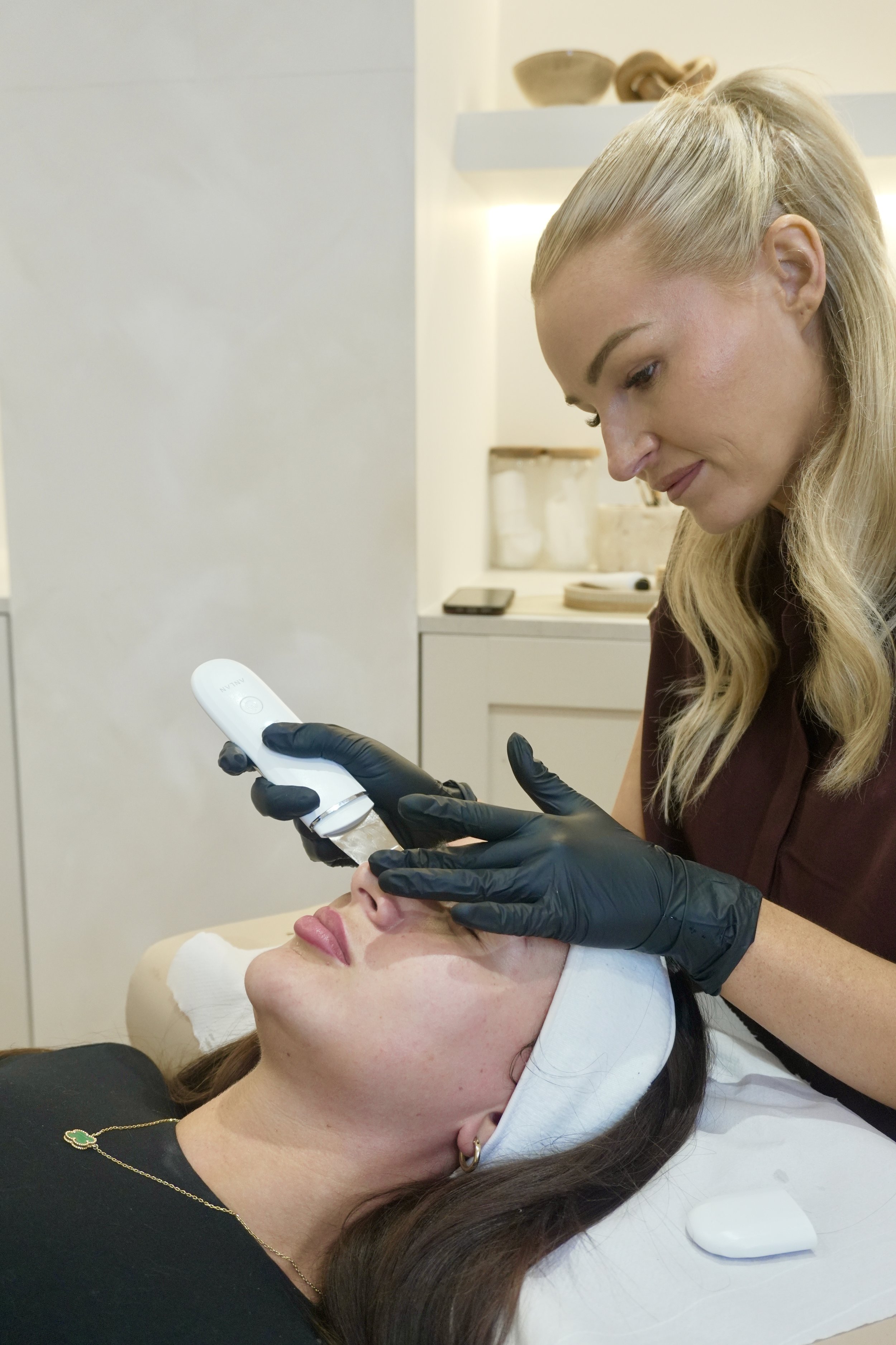 A woman receiving a facial treatment while lying on a bed with a white towel on her head, in a spa or clinic setting, with a medical professional gently applying a device to her face.