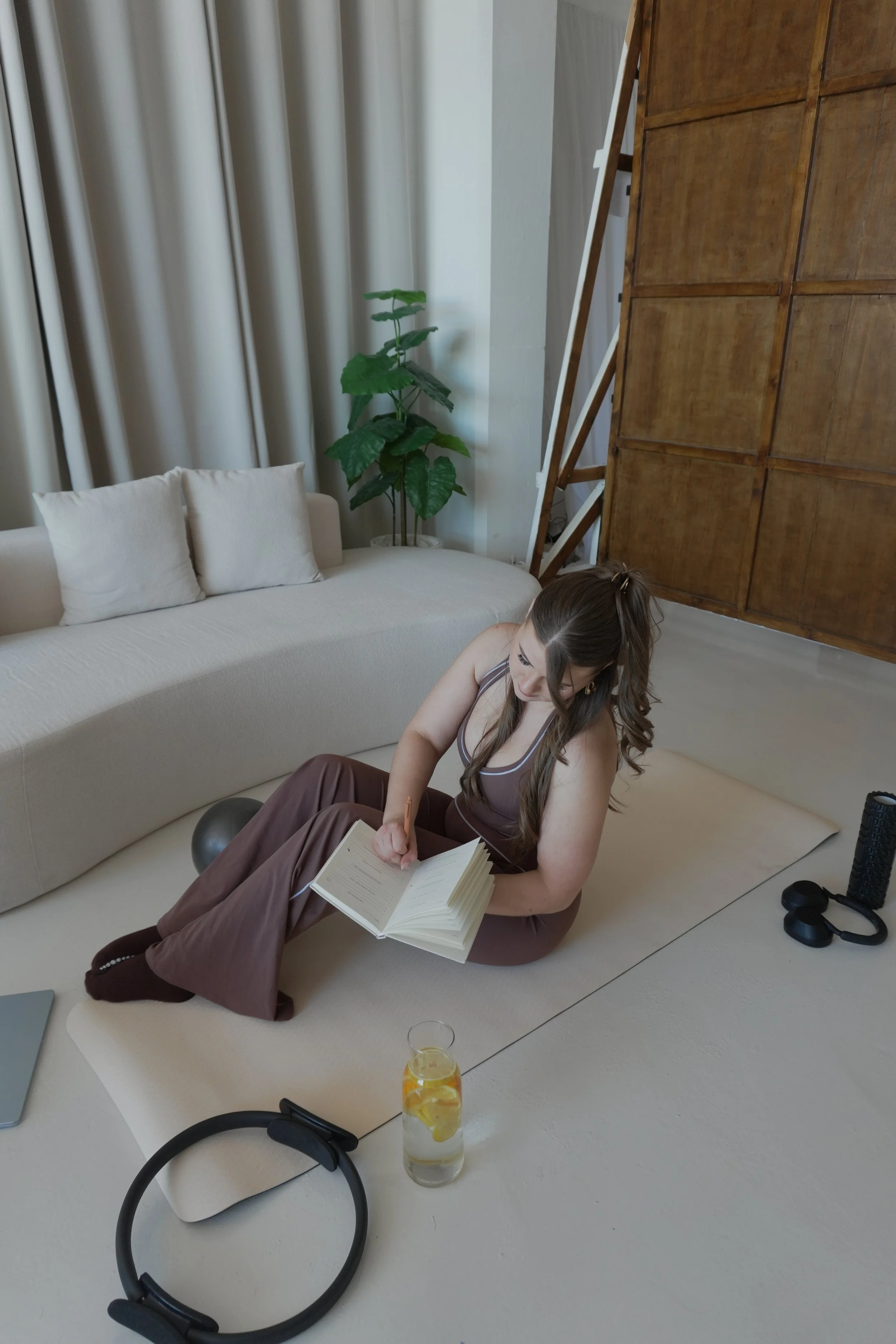 A woman with long brown hair in a ponytail is sitting on the floor, leaning against a beige couch, reading a book. There is a black dumbbell and a glass of orange-colored beverage with lemon slices on a tray in front of her. In the background, there 