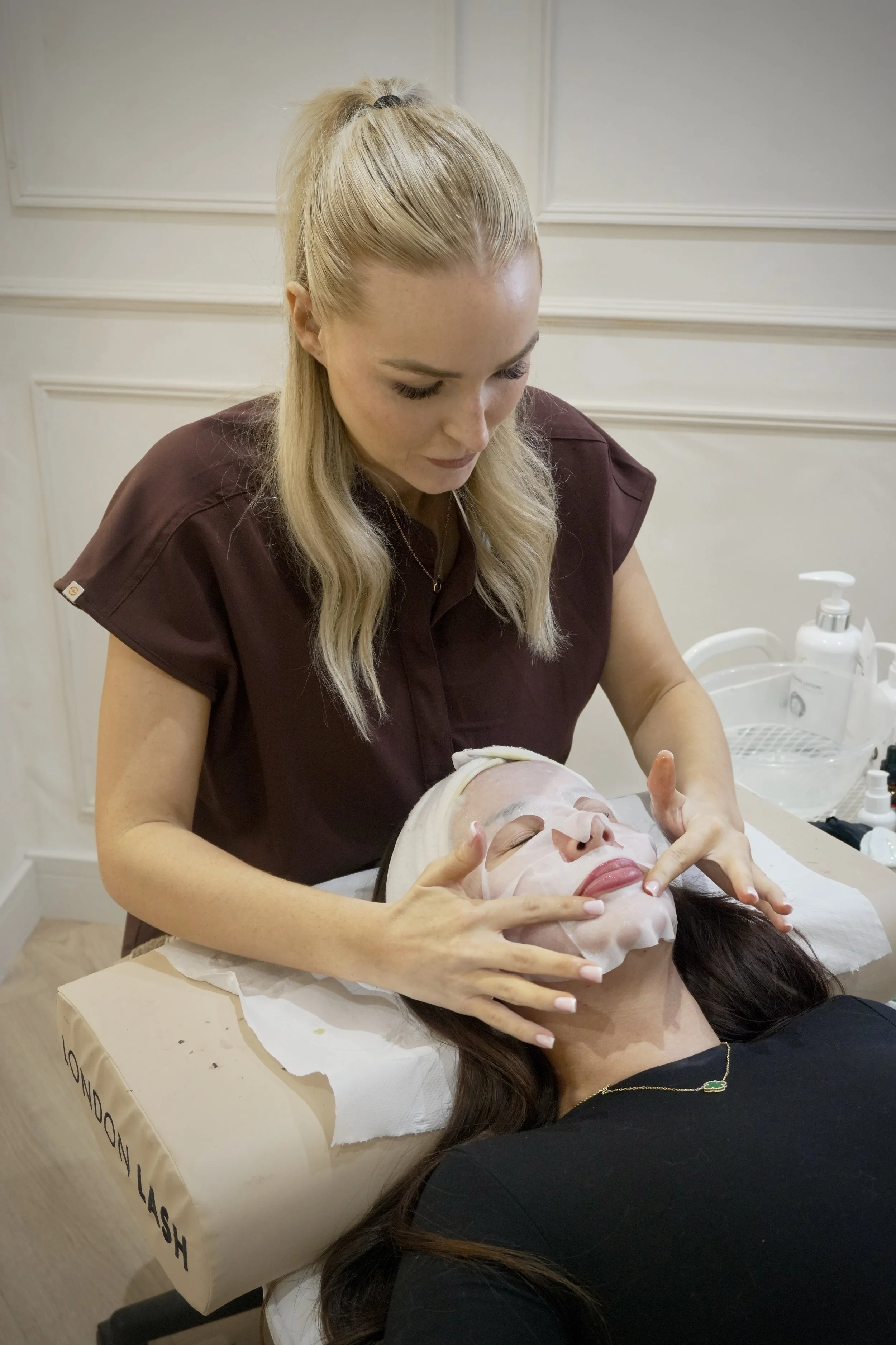 A woman with blonde hair receiving a facial treatment from a skincare professional, lying on a white treatment bed in a spa or clinic setting.