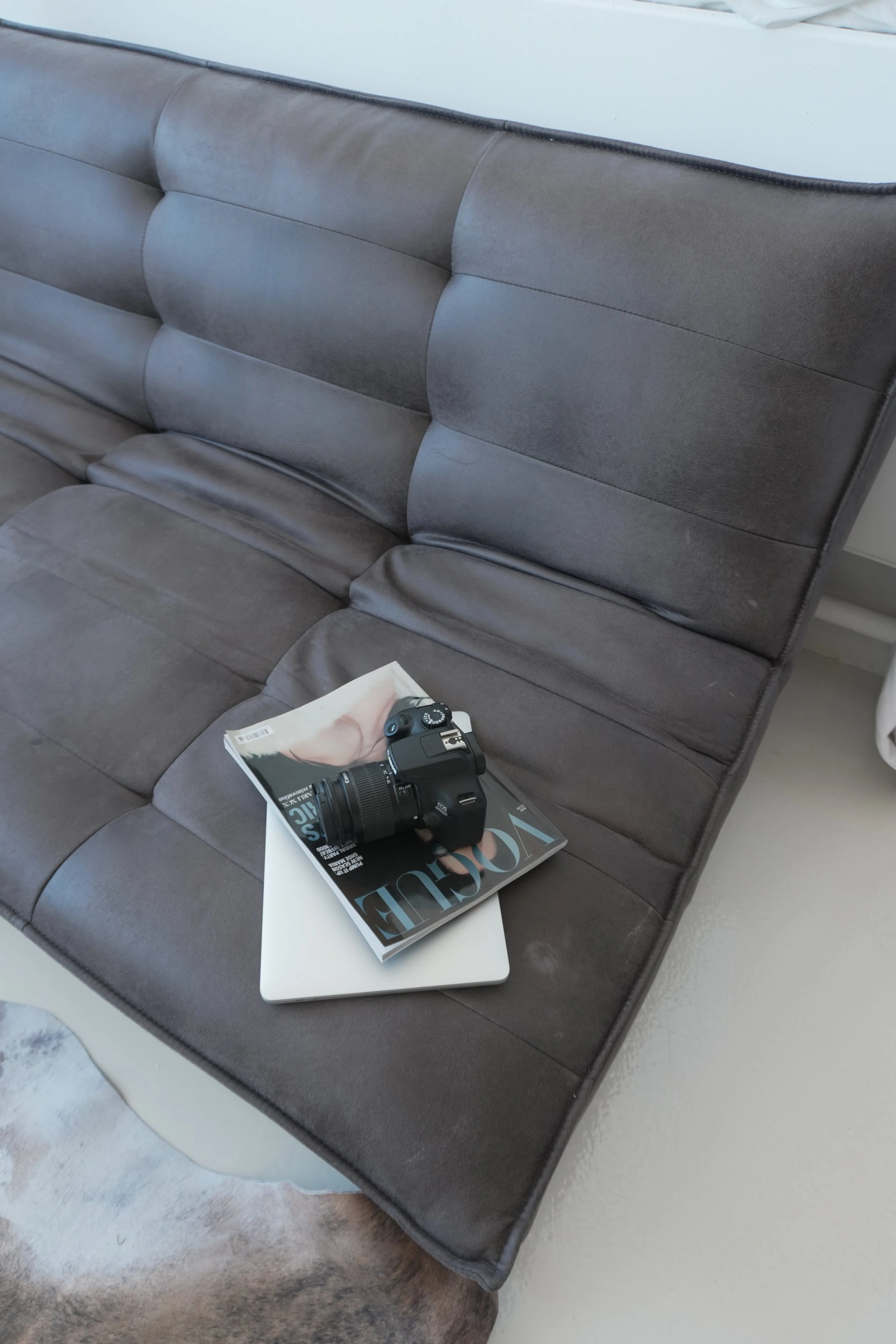 Gray upholstered couch with a magazine, a laptop, and a camera placed on it in a modern living space.
