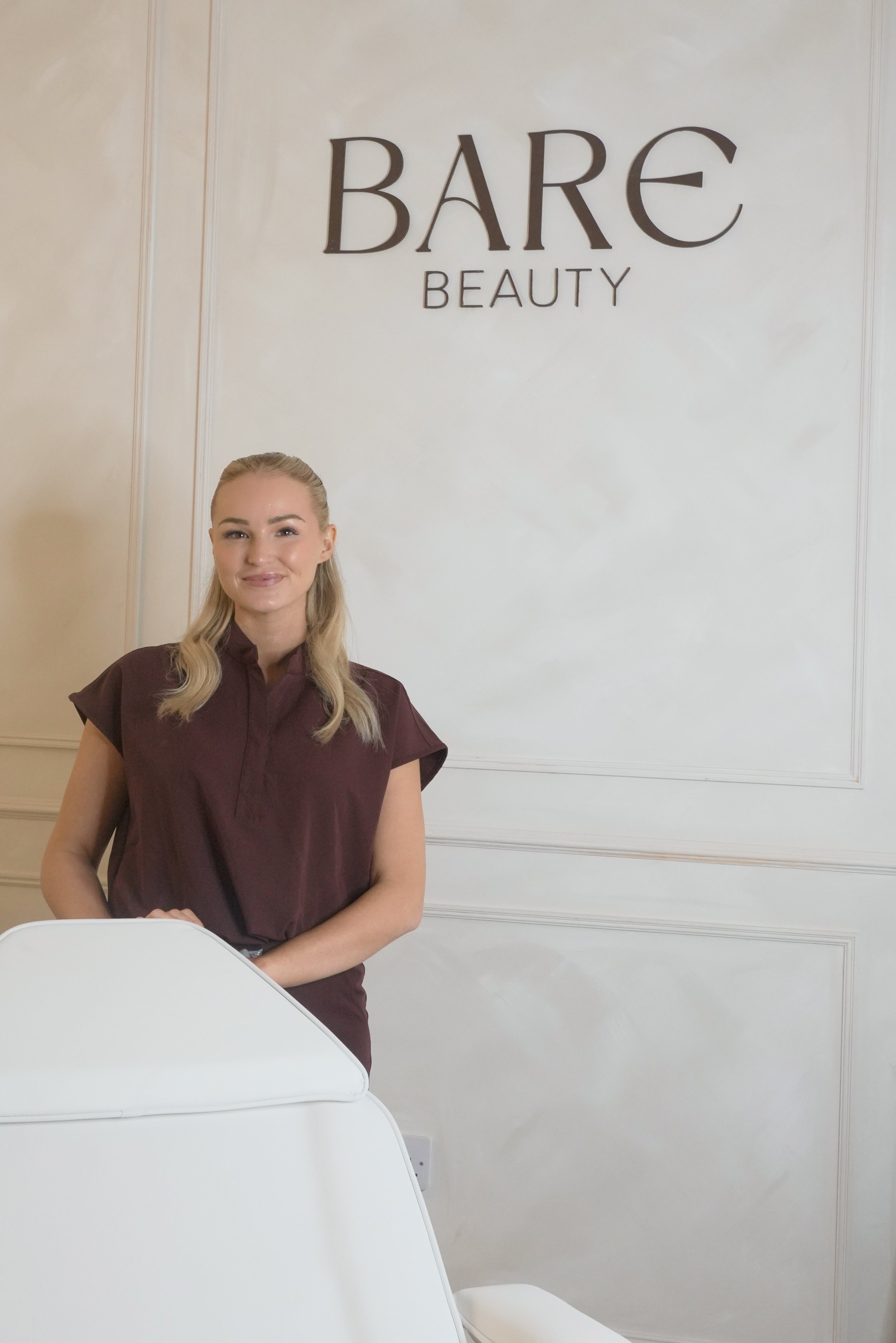 A woman with blonde hair in a bun, wearing a dark purple top, standing in front of a white wall with a sign that says "BARE BEAUTY."