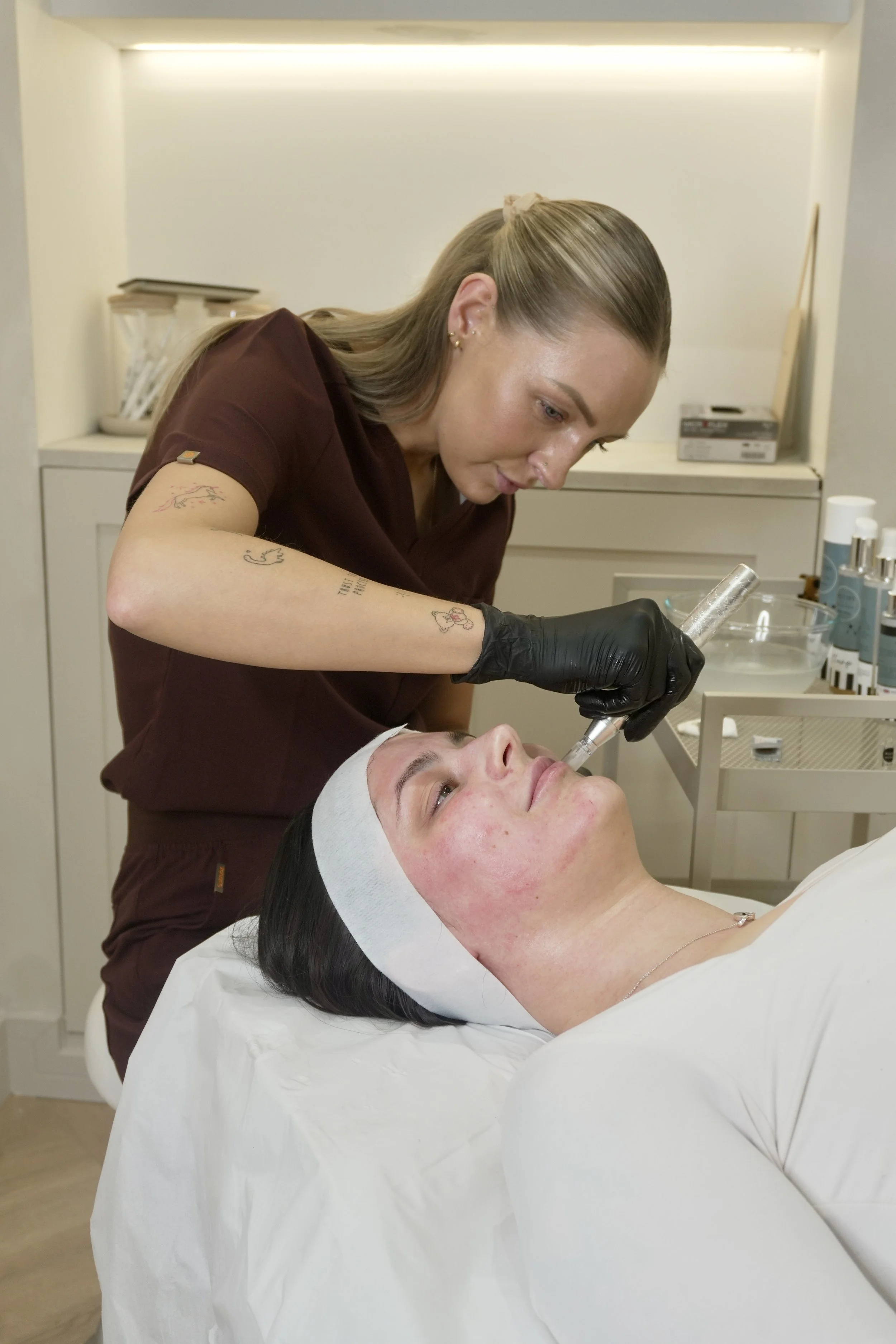 A woman lying on a medical bed with her eyes closed, receiving a cosmetic or dermatological treatment from a practitioner in a clinical setting.