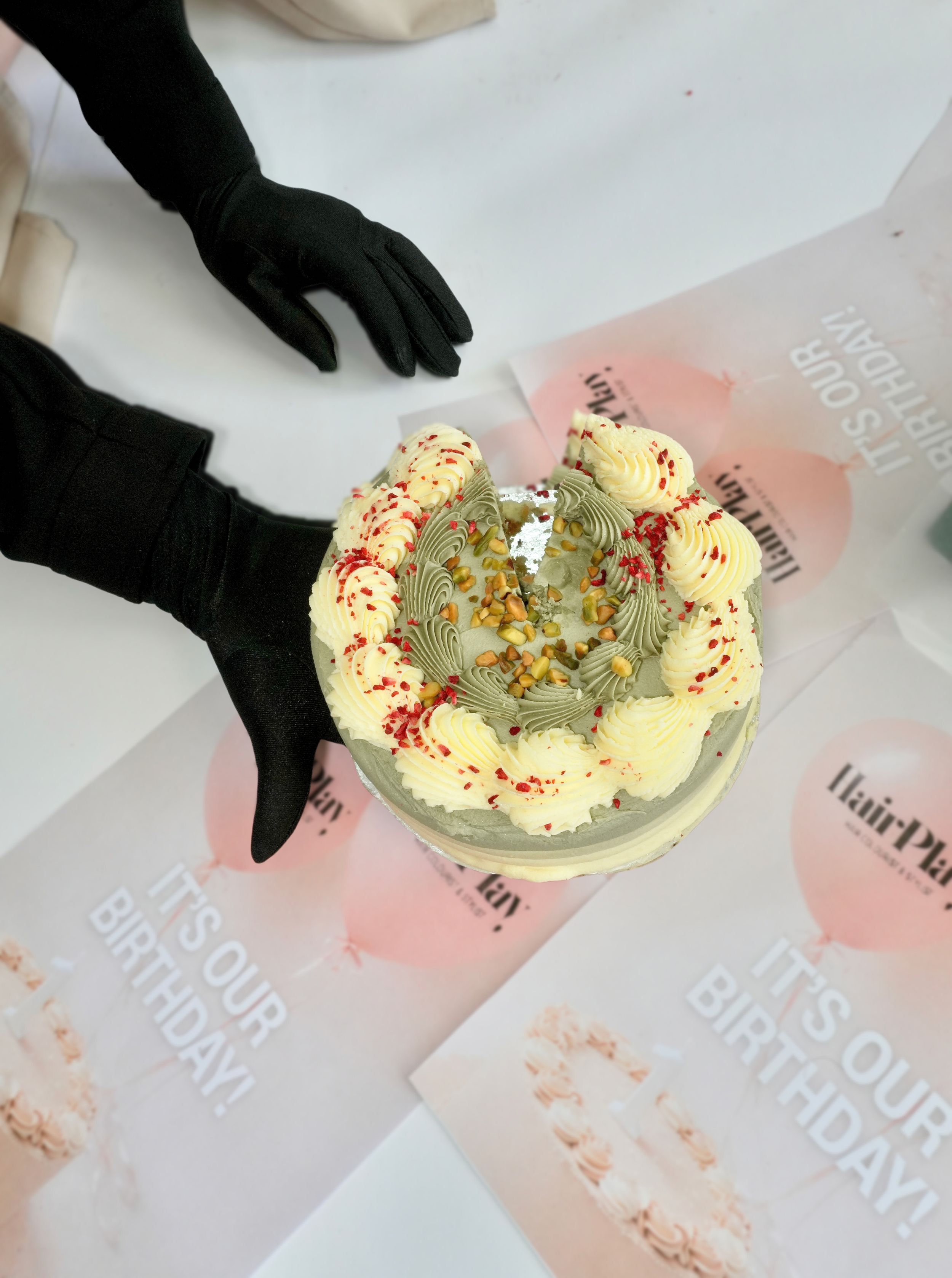 A birthday cake decorated with cream swirls, red sprinkles, and chopped nuts, held by someone wearing black gloves, on a table with a pink and white paper banner that reads 'It's Our Birthday!'