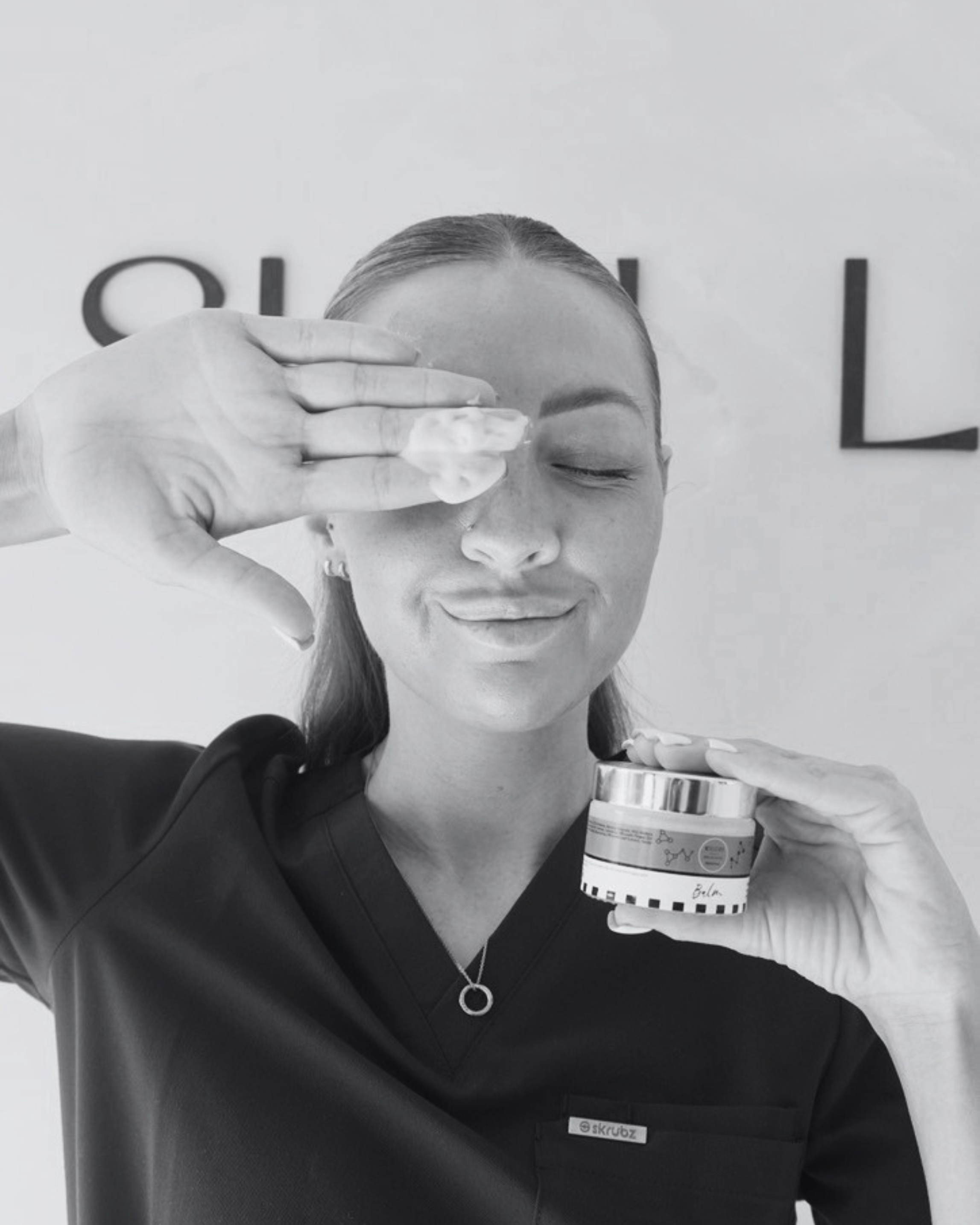 A woman with pulled back hair is holding a can of whipped cream in front of her face, with her eyes closed. She is wearing a dark shirt and a necklace with a circular pendant. The background has a partially visible word, possibly 'SKRUBZ'.