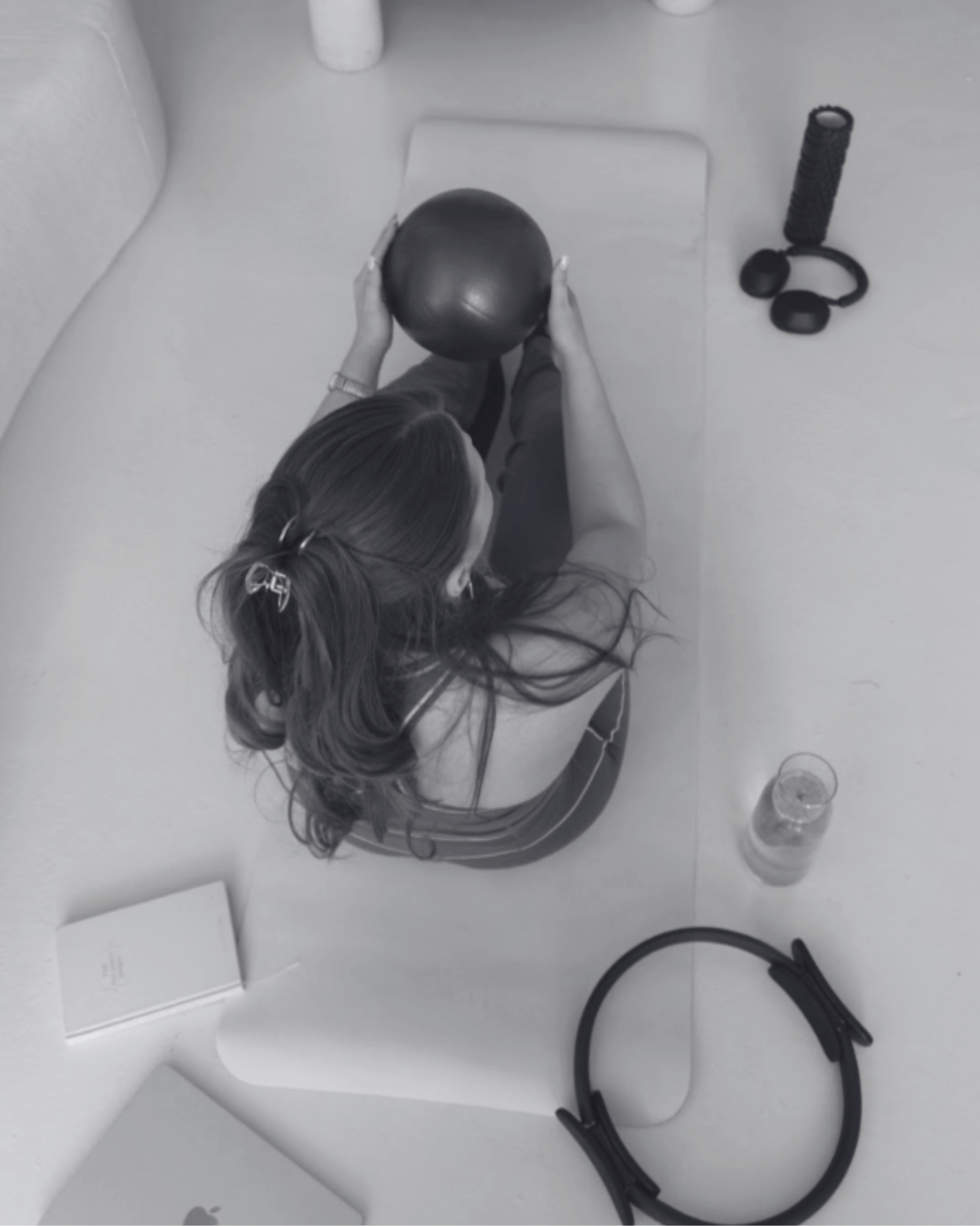 A woman with long dark hair is sitting on the floor holding a black globe. Nearby are various objects including a pair of black headphones, a white tablet, a white box, a transparent plastic container, a coiled black cable, and a black headband, all on a white surface.