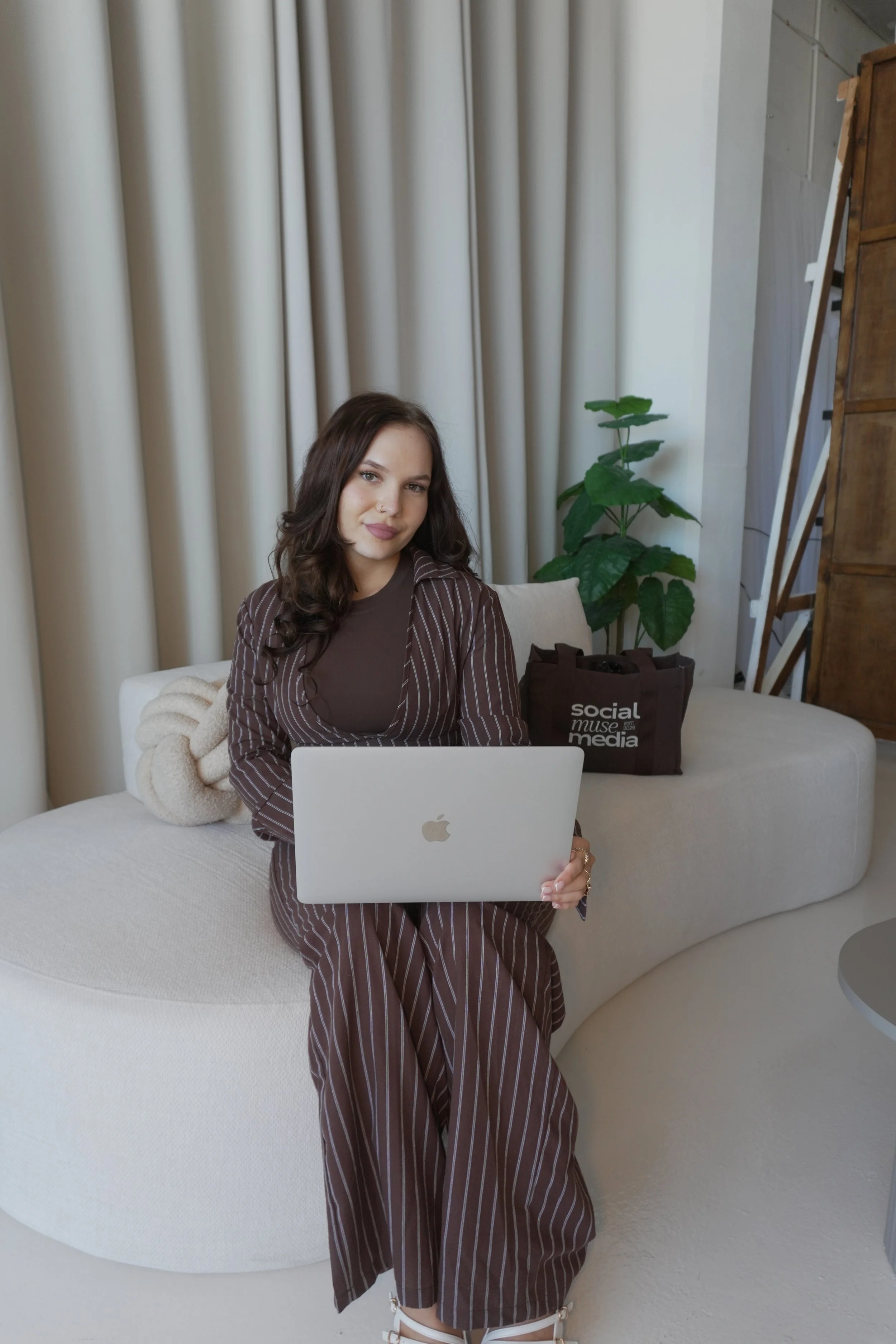 A woman with brown hair sitting on a white sofa, working on a silver MacBook. She is wearing a brown striped suit and white high-heeled shoes. Behind her, there are cream-colored curtains, a green plant, and a brown bag with the text 'social media' on it.