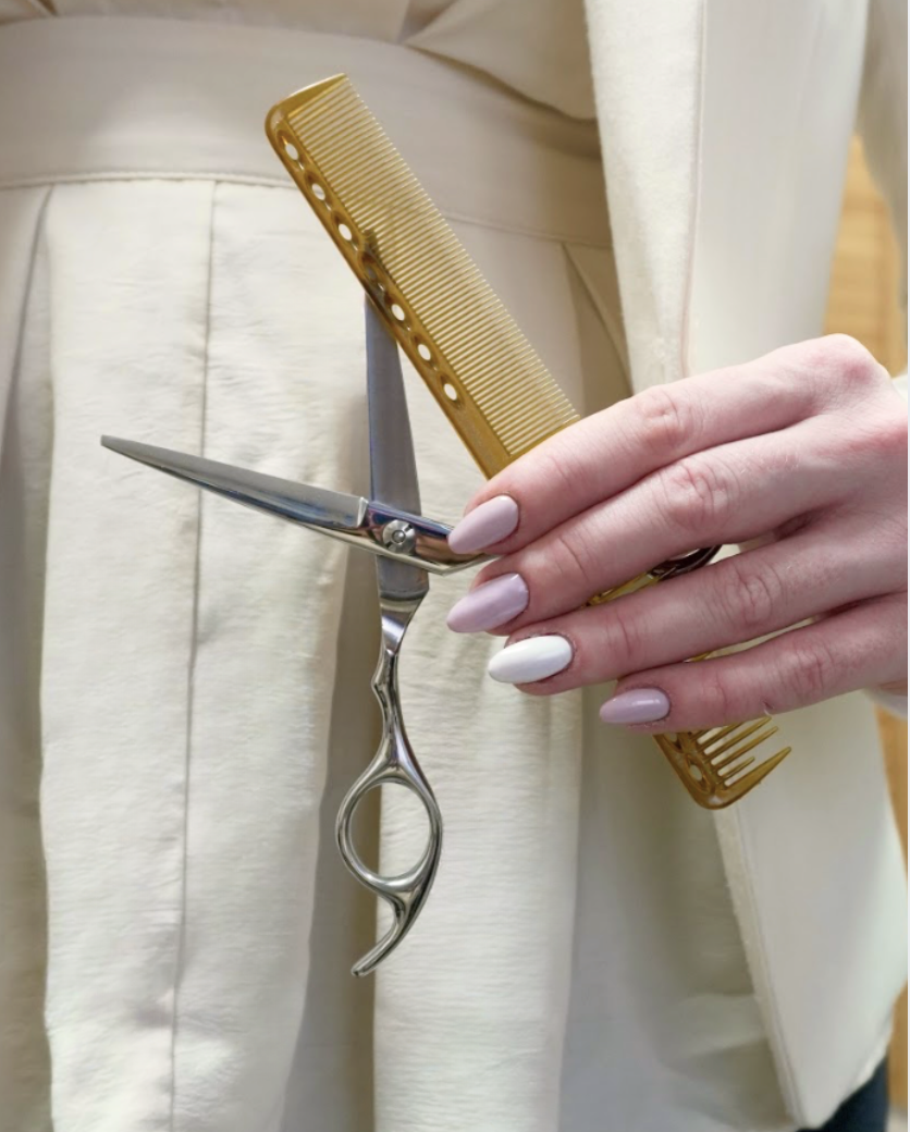 A person holding a pair of scissors, a yellow comb, and a light pink nail polish. The person is wearing a white or cream-colored coat or jacket.