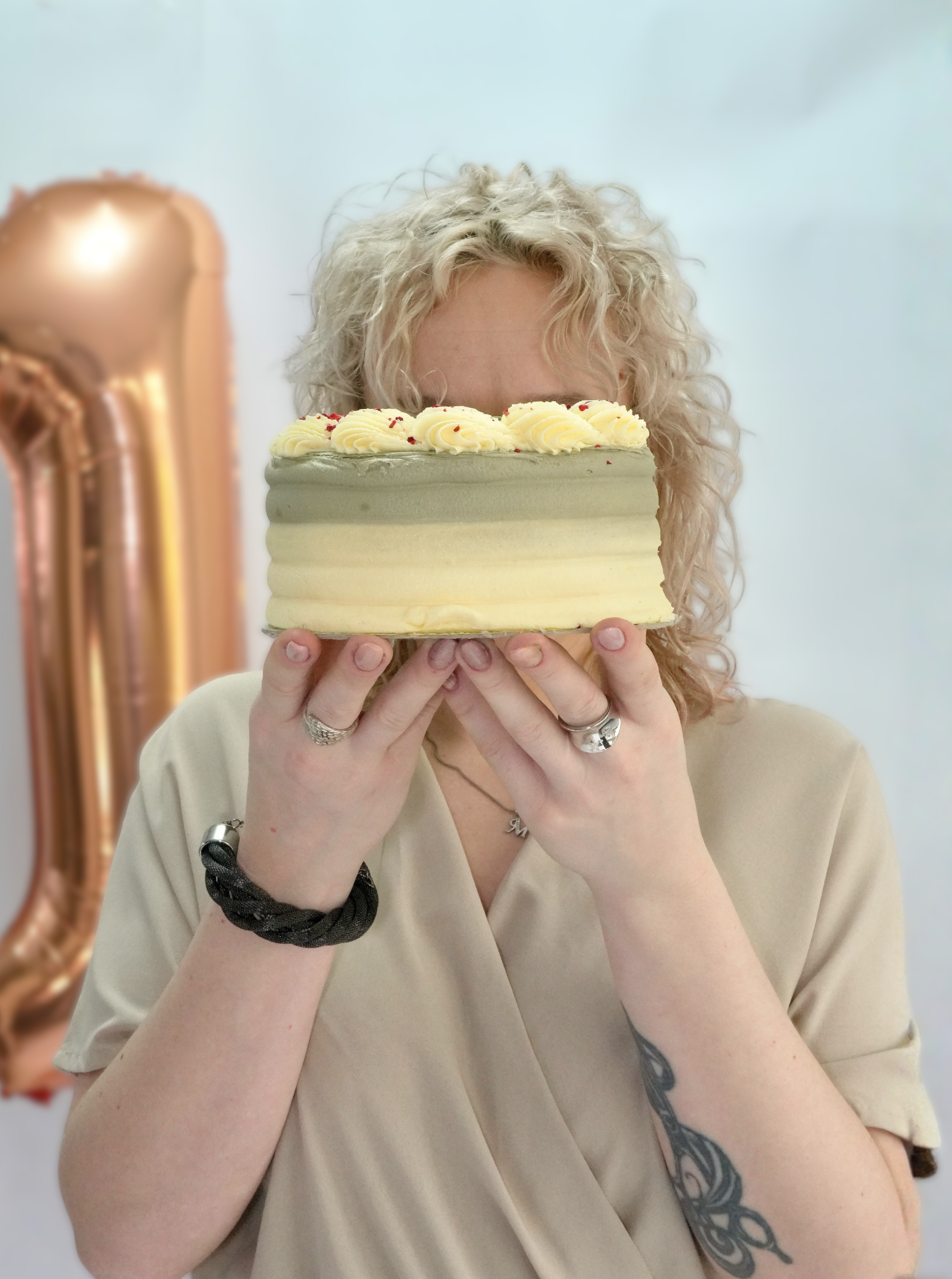 A woman with curly blonde hair holding a birthday cake with white frosting and yellow icing swirls, partially hiding her face.