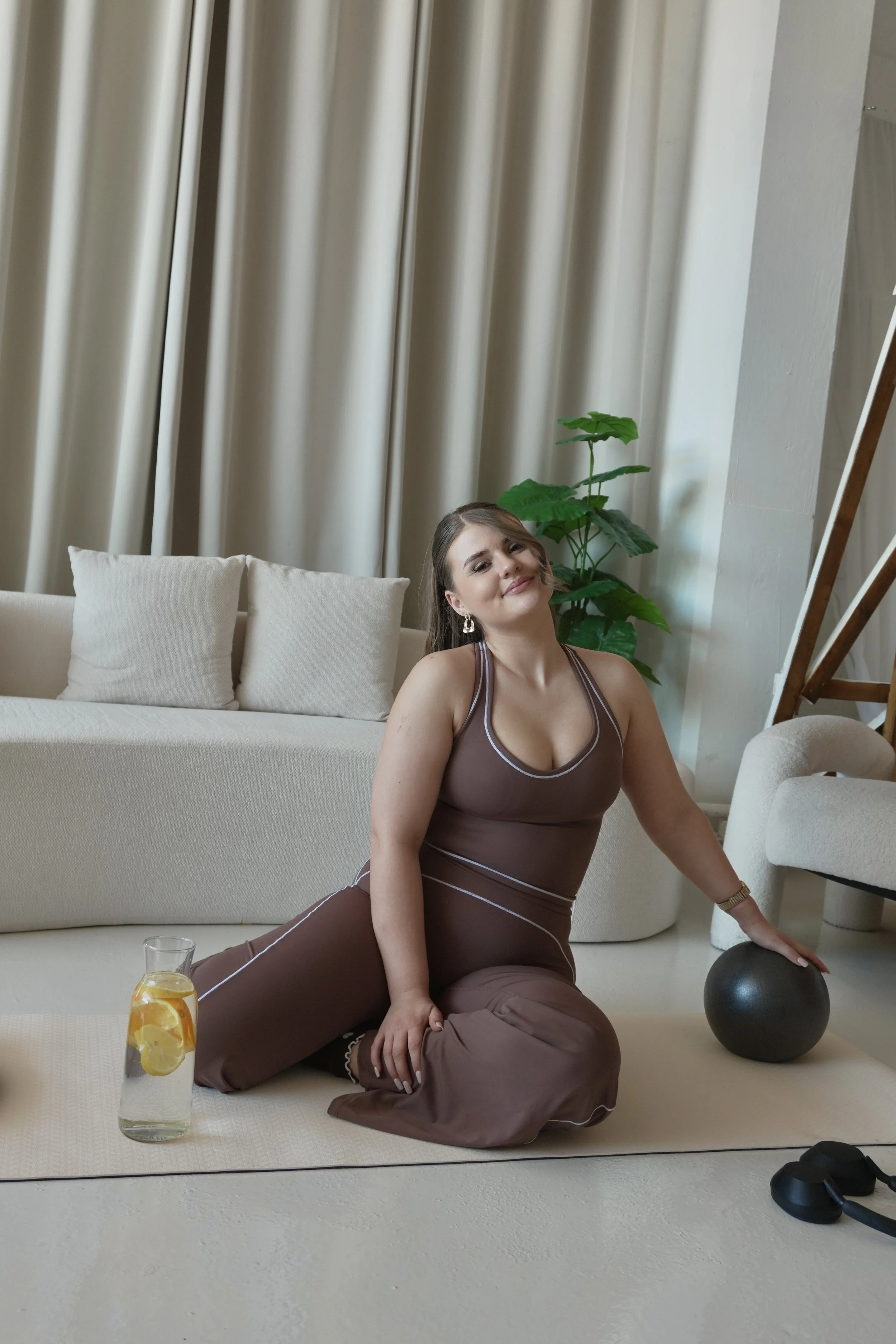 A young woman with brown hair and earrings, wearing a brown athletic outfit, is sitting on a beige carpet. She is smiling and looking at the camera with her left hand on a black exercise ball. There are beige pillows on a sofa behind her, a green pla