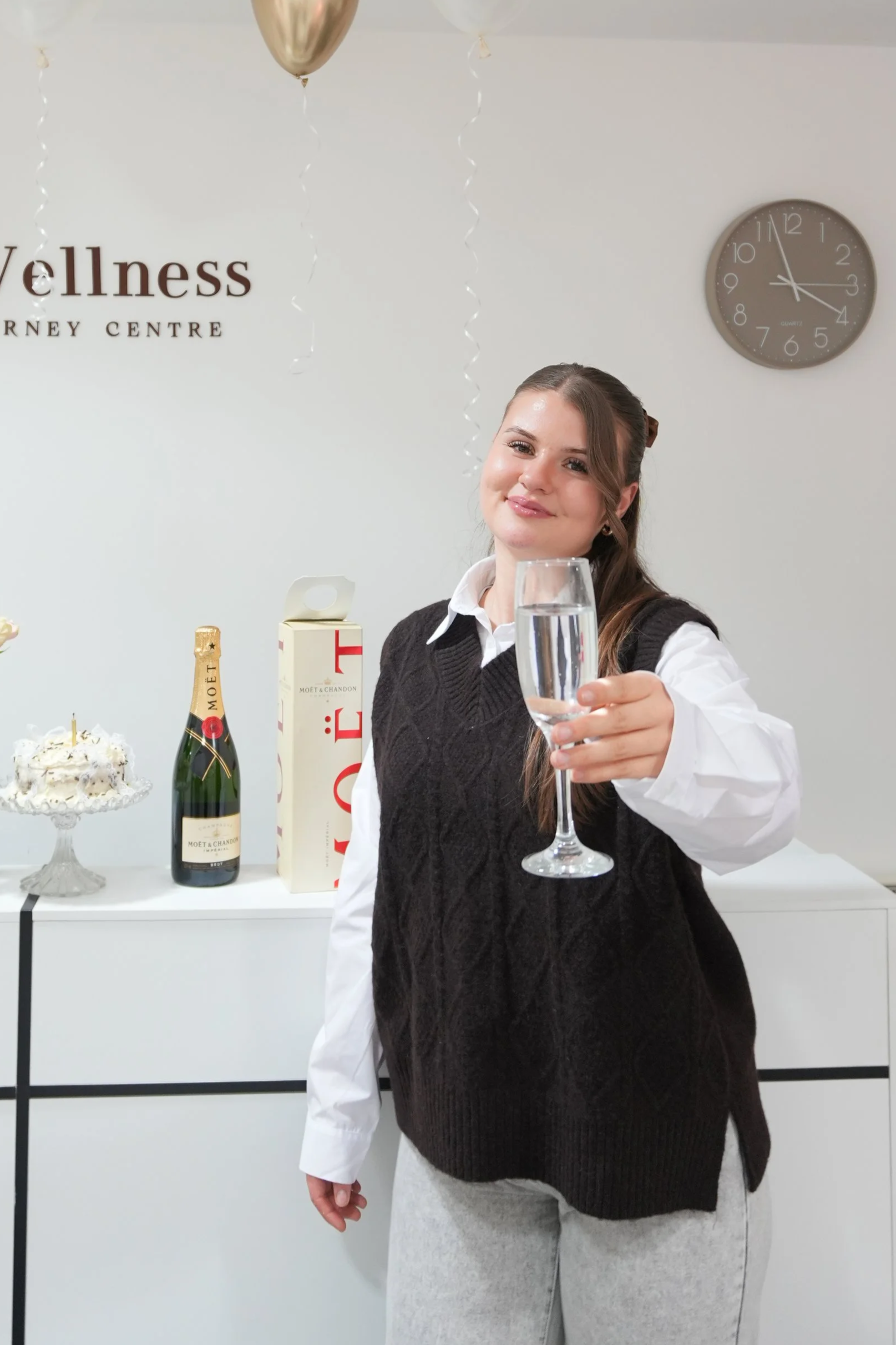 Young woman at celebration holding a glass of champagne, standing in front of a table with champagne bottles, a cake, and a gift box, with a wall clock and partial sign in the background.