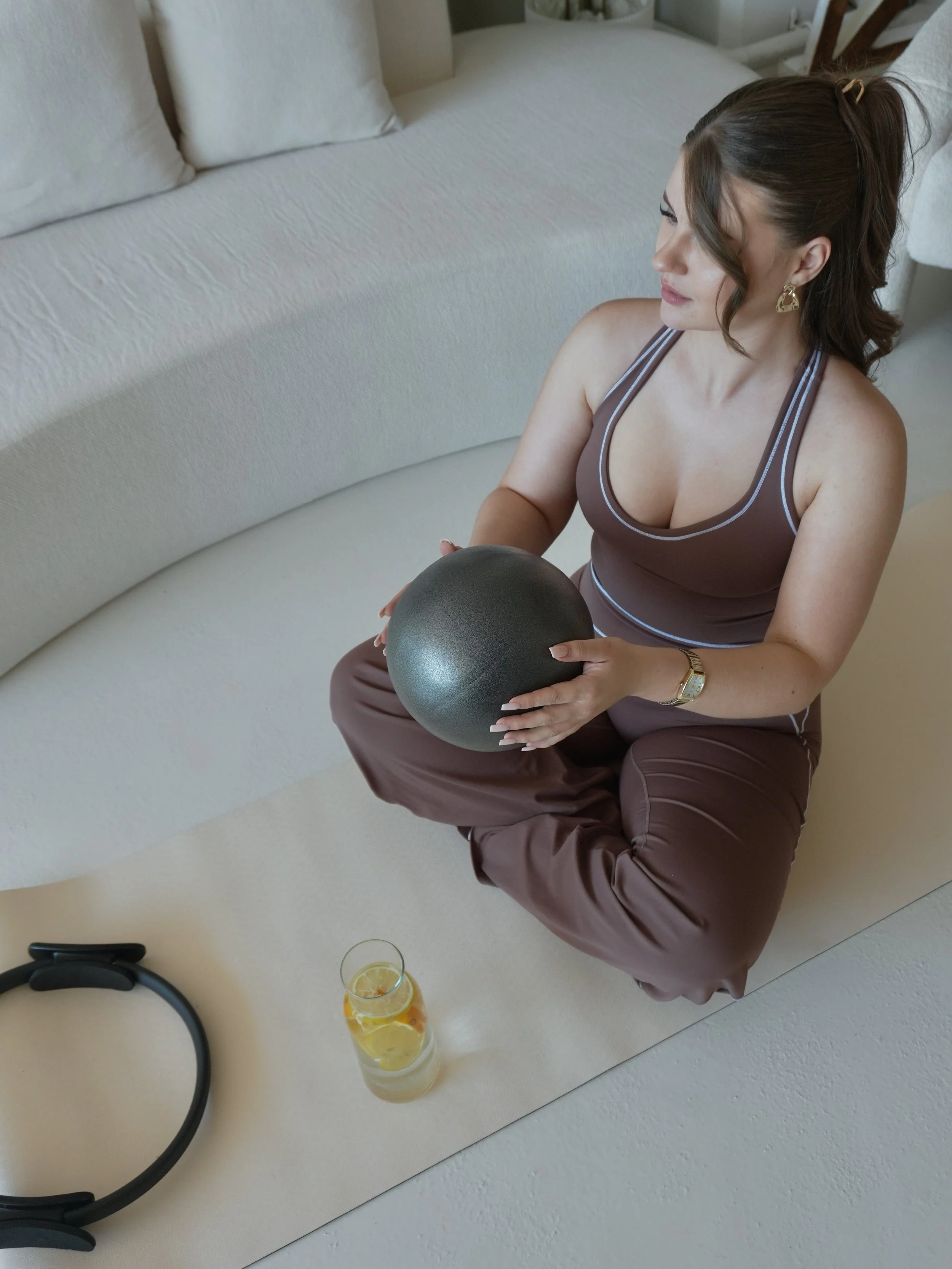 A woman sitting on the floor holding a black kettlebell, with two glasses of lemon and orange flavored drink nearby, on a white mat in a room with a white couch.