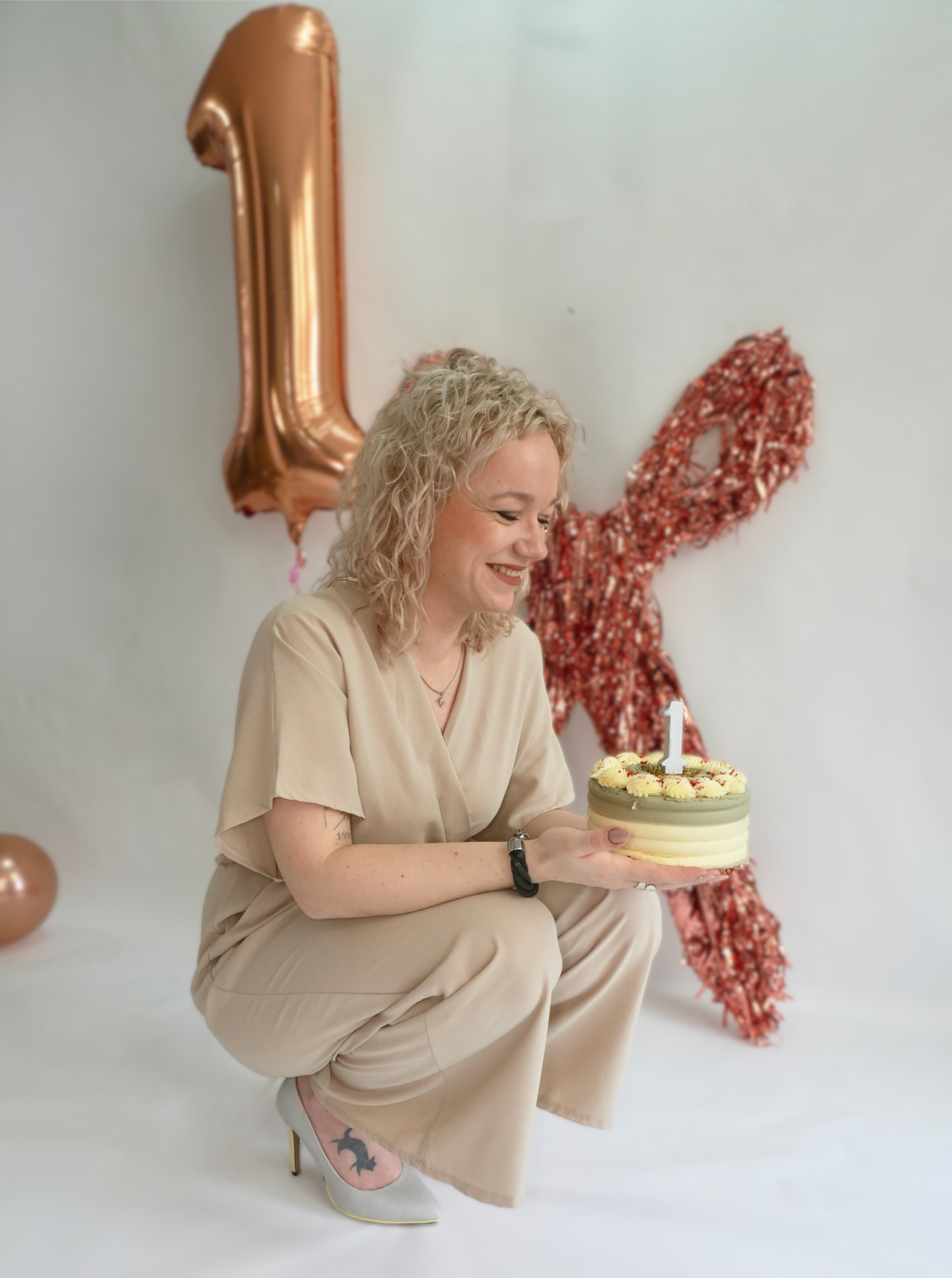 A woman with curly blonde hair smiling while sitting on her heels, holding a birthday cake with a single lit candle, in front of a backdrop with metallic balloons and a red, glittery number one balloon.