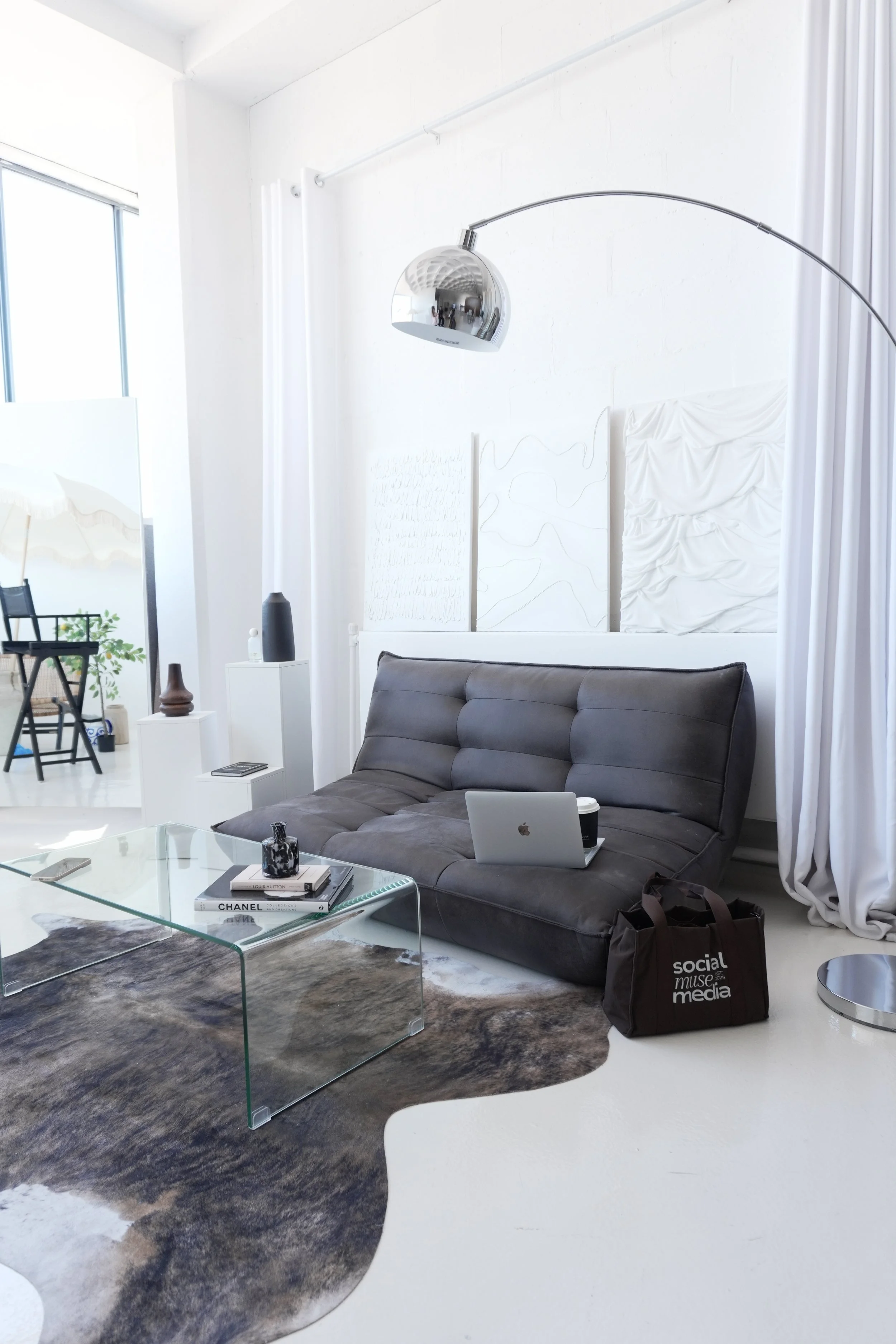 Modern living room with a black leather sofa, glass coffee table with books and a decorative item, patterned rug, and artwork on the white wall. There is a laptop and a to-go coffee cup on the sofa, a black and white bag on the floor, and a corner with a small table and bookshelf. The space is bright with large windows and white walls.