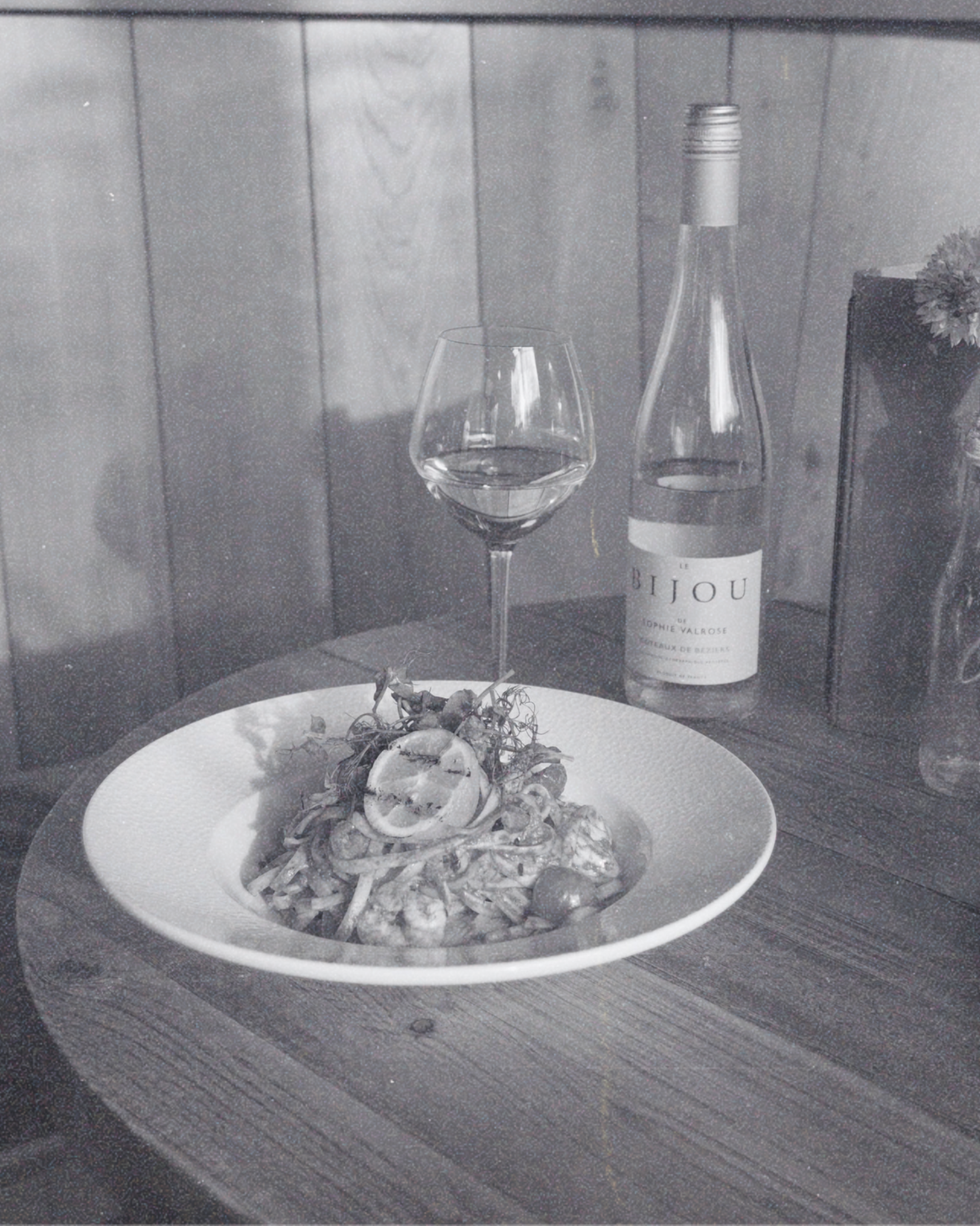 A glass of wine, a bottle of wine, and a plate of pasta on a table with a wooden paneled wall in the background.