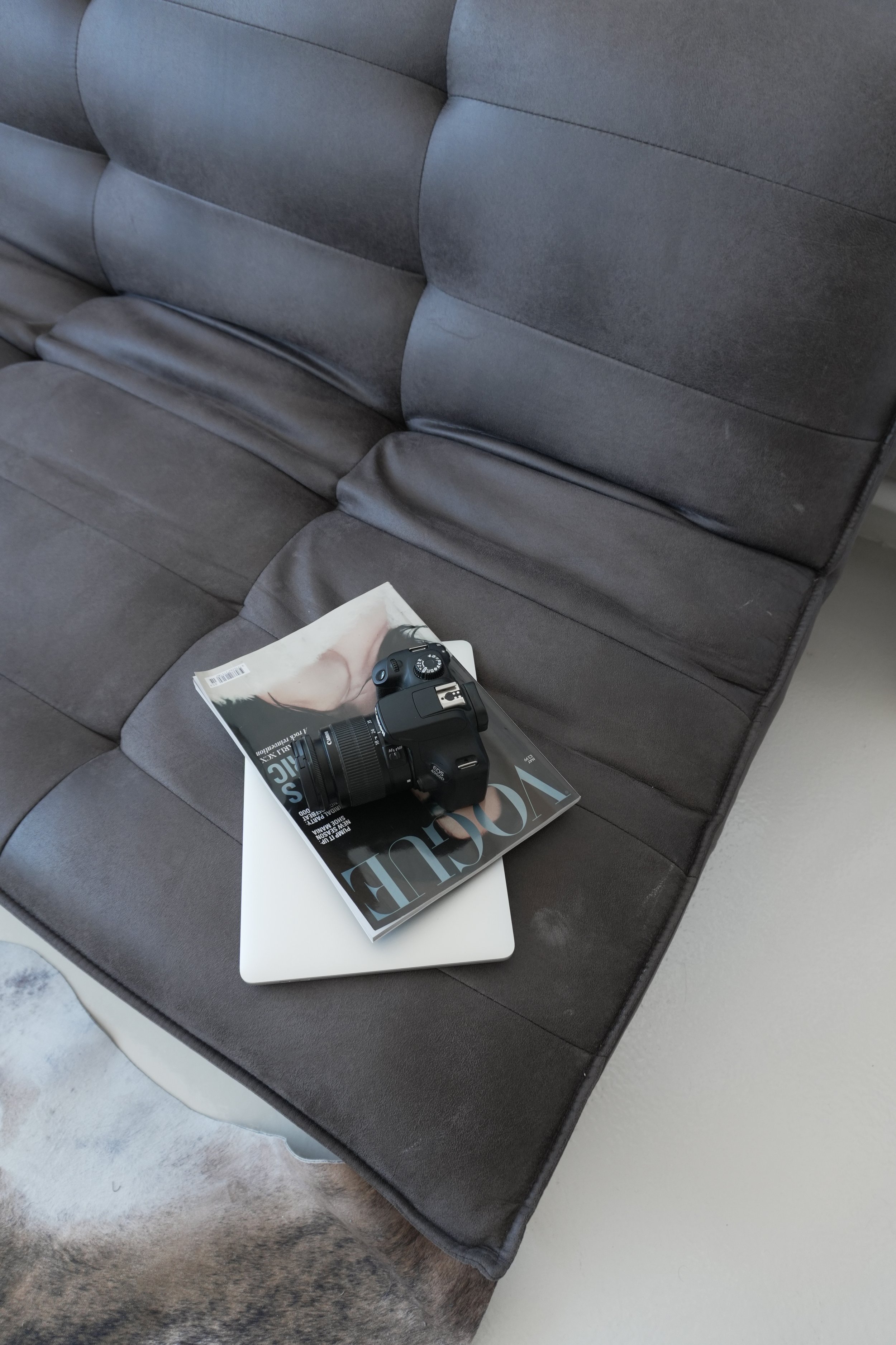 A dark gray couch with a magazine, a camera, and a closed white laptop on it. The magazine features a model on the cover, and the camera is a Canon DSLR.