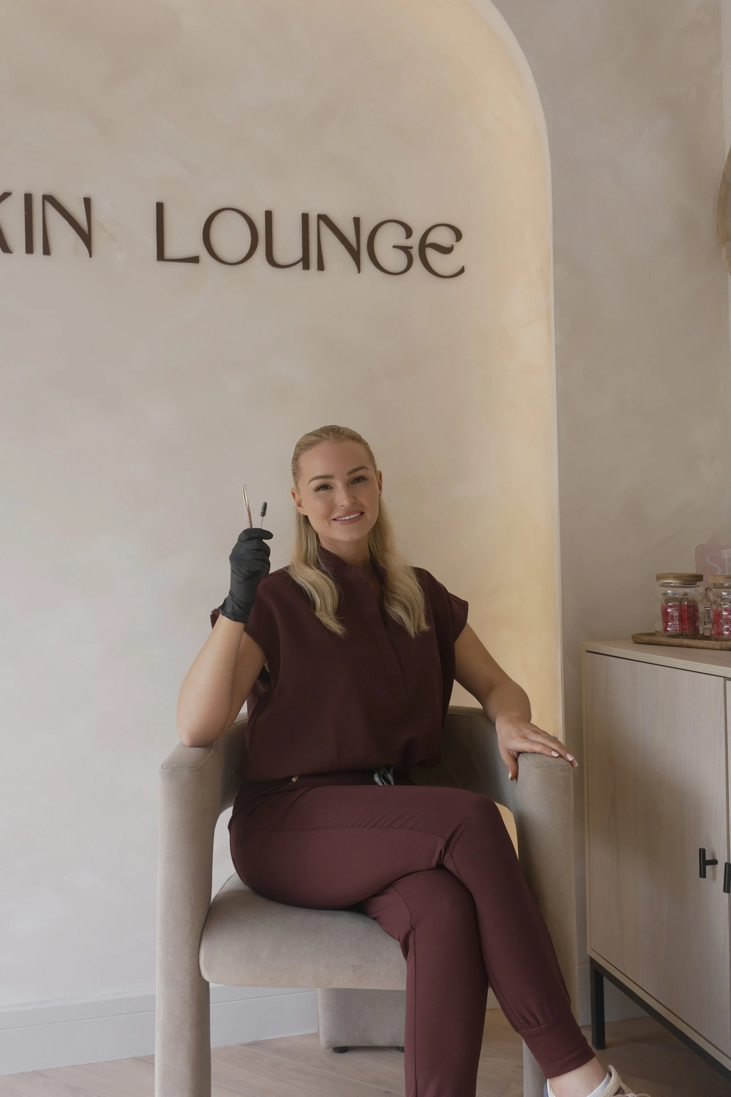 A woman sitting in a beige armchair in a salon or spa, smiling and holding a pair of scissors and a comb, with the sign 'KAIN LOUNGE' on the wall behind her.