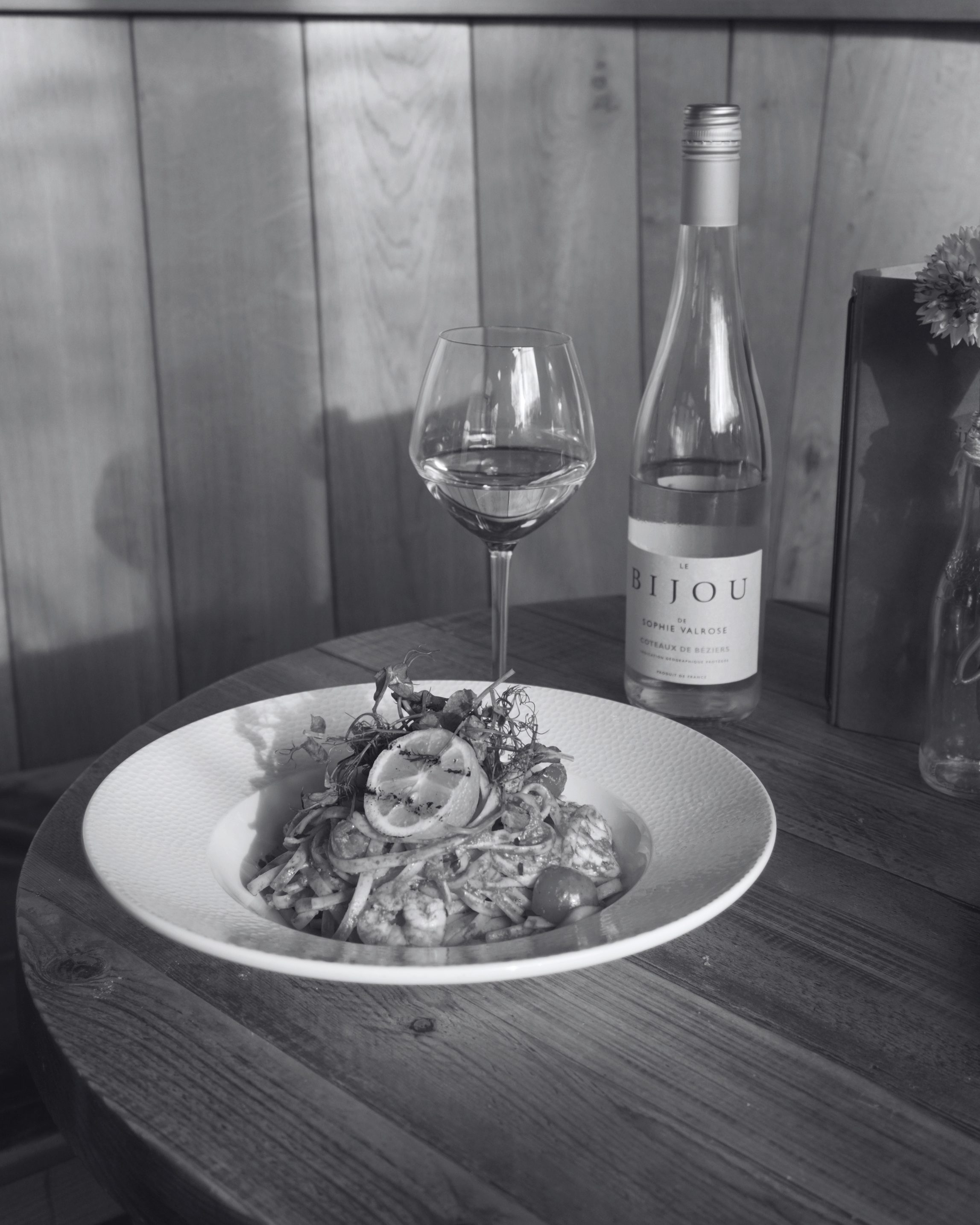 A plate of pasta with sliced lemons and herbs, a glass of water, and a bottle of wine on a wooden table, with a wooden-paneled wall in the background.