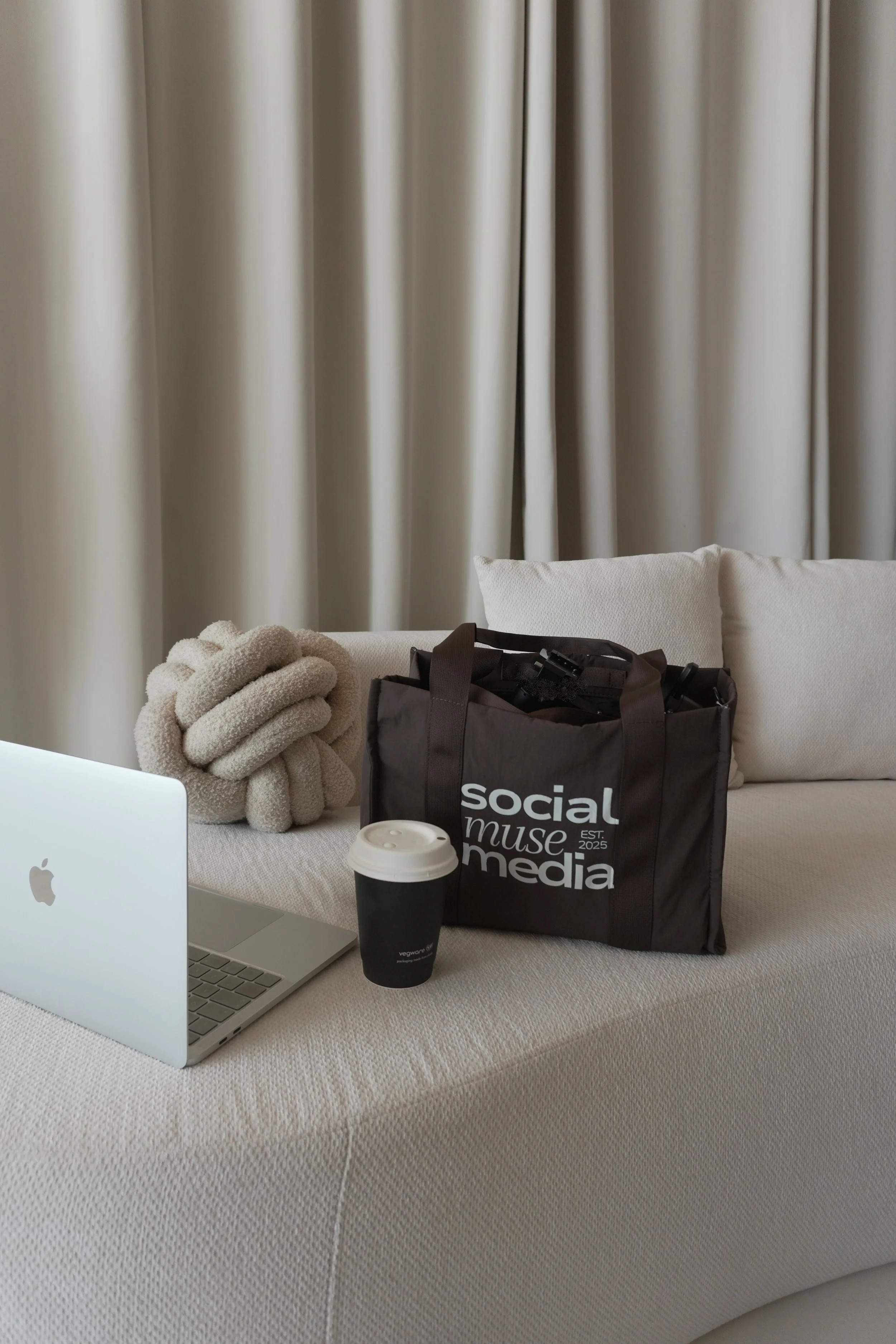 A beige fabric couch with a beige blanket, a black tote bag with white text, a white laptop, and a black to-go coffee cup placed on it. Behind the couch, beige curtains are partially visible.