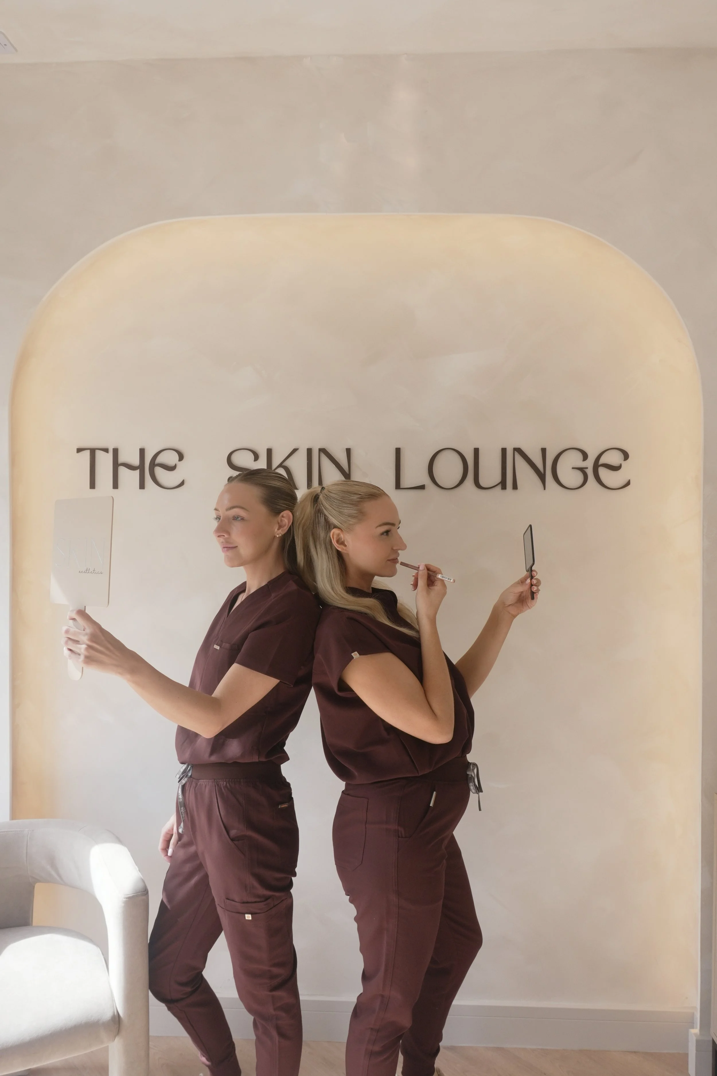 Two women in matching maroon scrubs stand back to back, each holding a mirror and looking into the distance. They are in a room with a neutral beige wall that has the words "THE SKIN LOUNGE" on it.