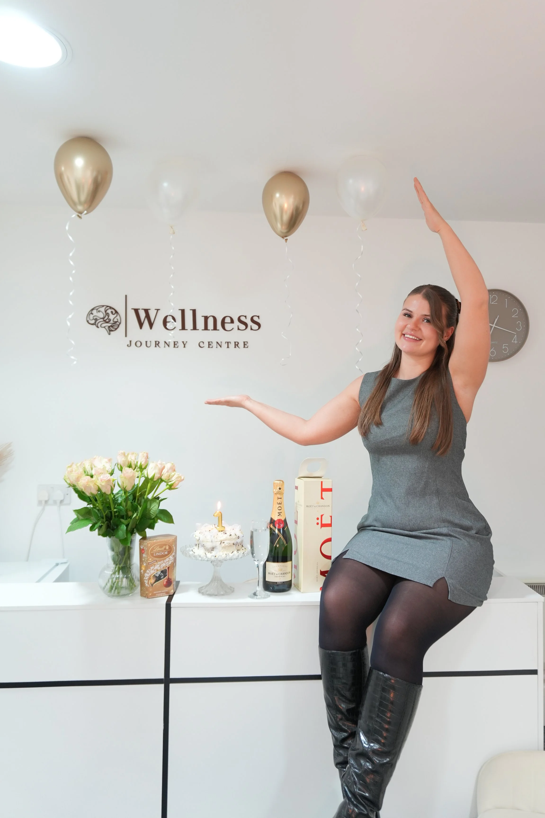 A woman dressed in a gray sleeveless dress and black thigh-high boots sitting on a white table in a wellness center. She is smiling and raising one arm, with her other arm extended. On the table are a bouquet of pink roses, a birthday cake with a candle, a bottle of Moët & Chandon champagne, and various gift boxes. The background includes a wall with balloons, a wellness center sign, and a clock.