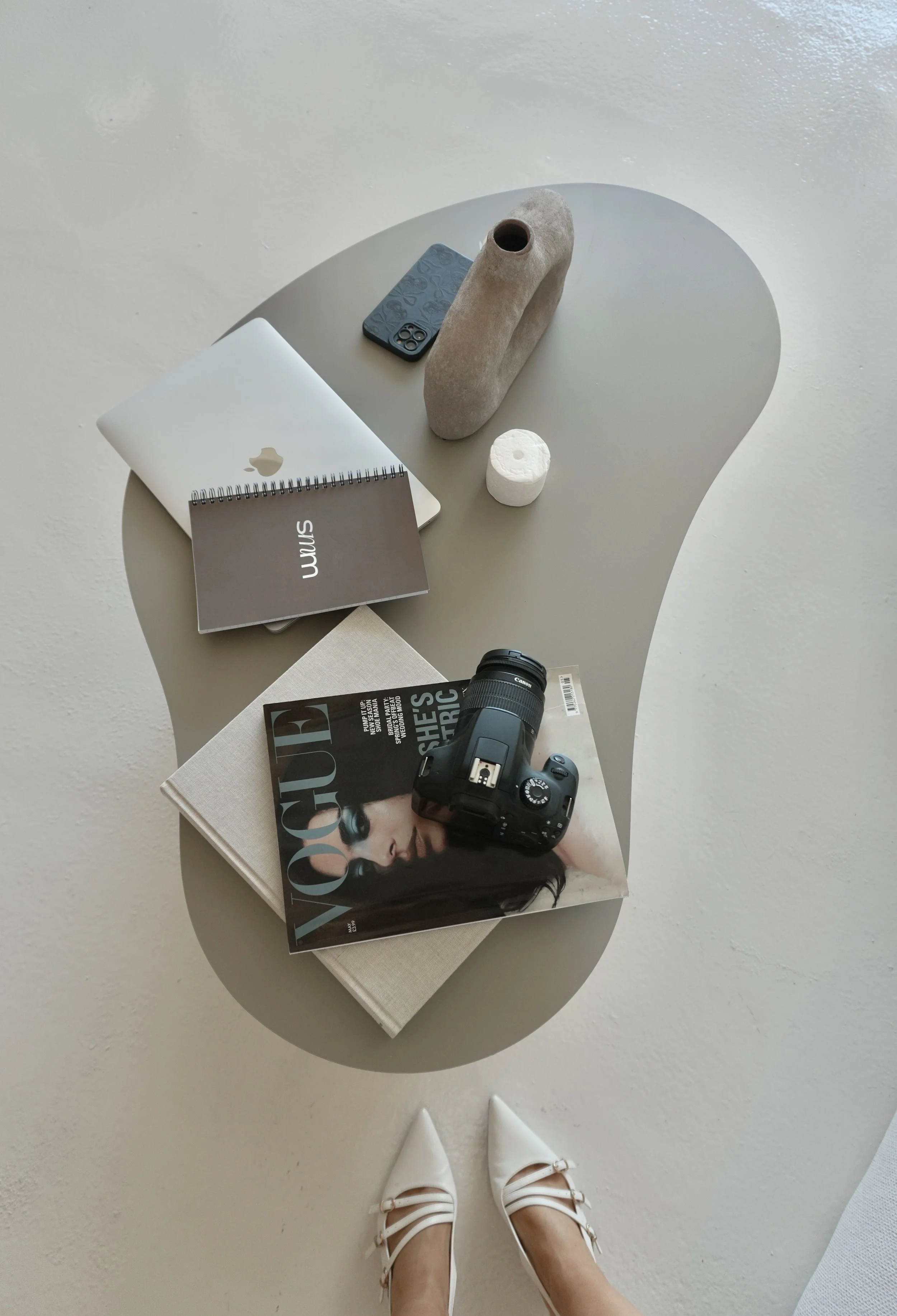 A person's feet wearing white shoes with bows are seen standing on a light-colored floor, with a grey curved table holding various items including a closed laptop, a black camera, magazines, a small foam object, a tablet, and a decorative sculpture.