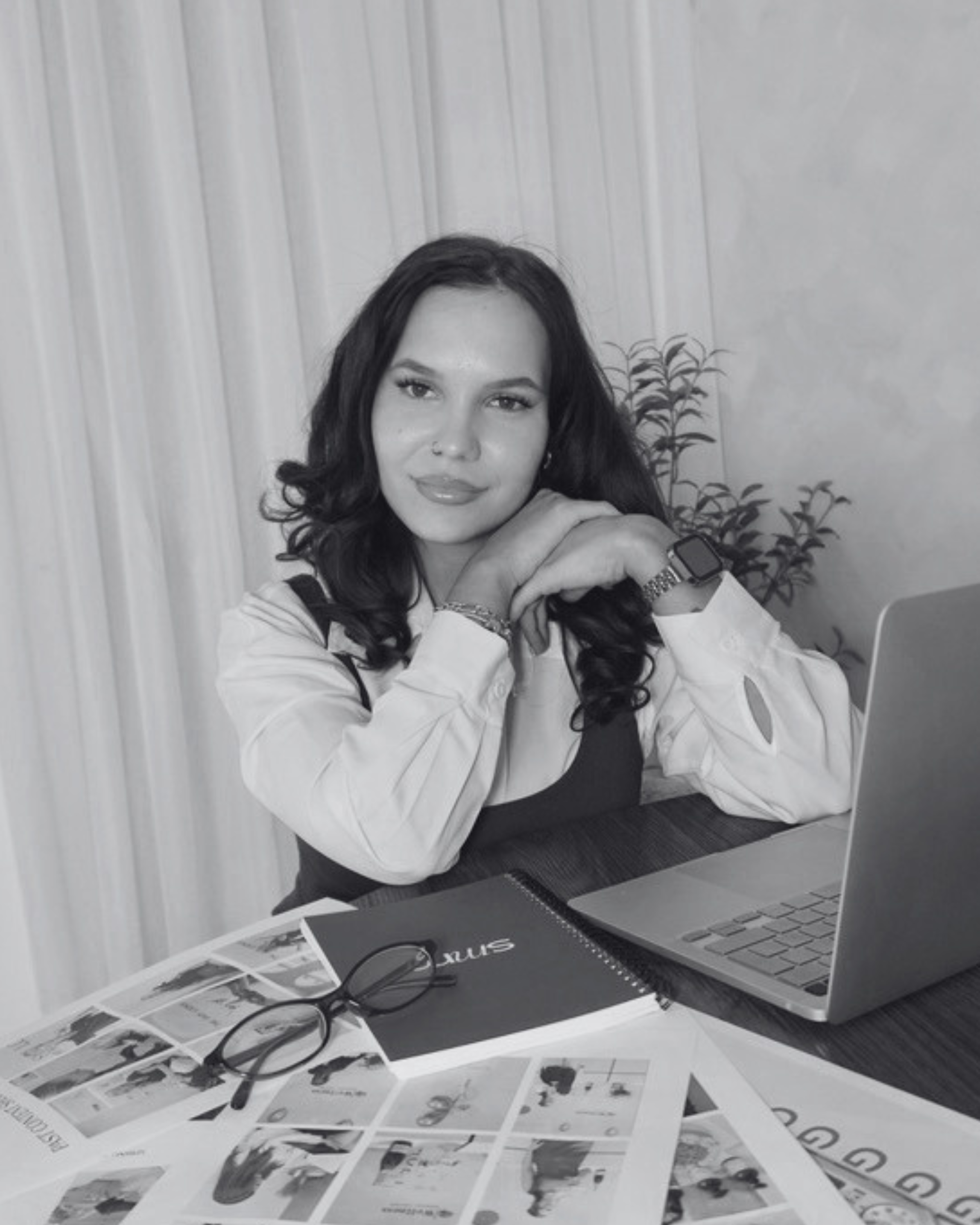 A woman with dark, wavy hair and a confident expression, sitting at a desk with a laptop, scattered printed photographs, a notebook, and eyeglasses, in black and white.
