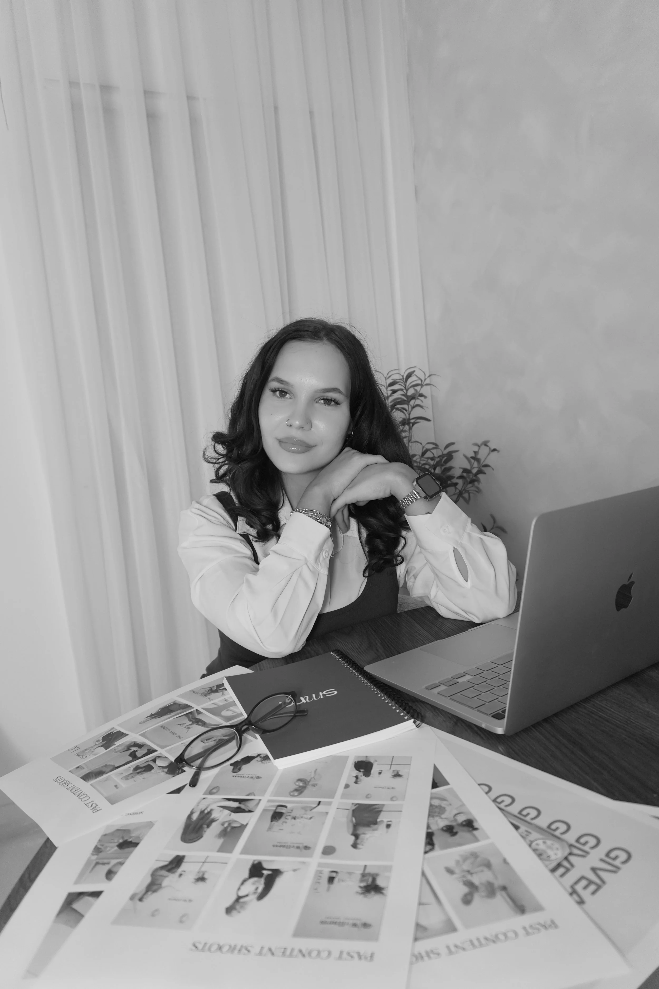 A young woman with dark wavy hair and a nose piercing sitting at a desk with a laptop, magazines, glasses, and sunglasses, in a room with white curtains and potted plants.