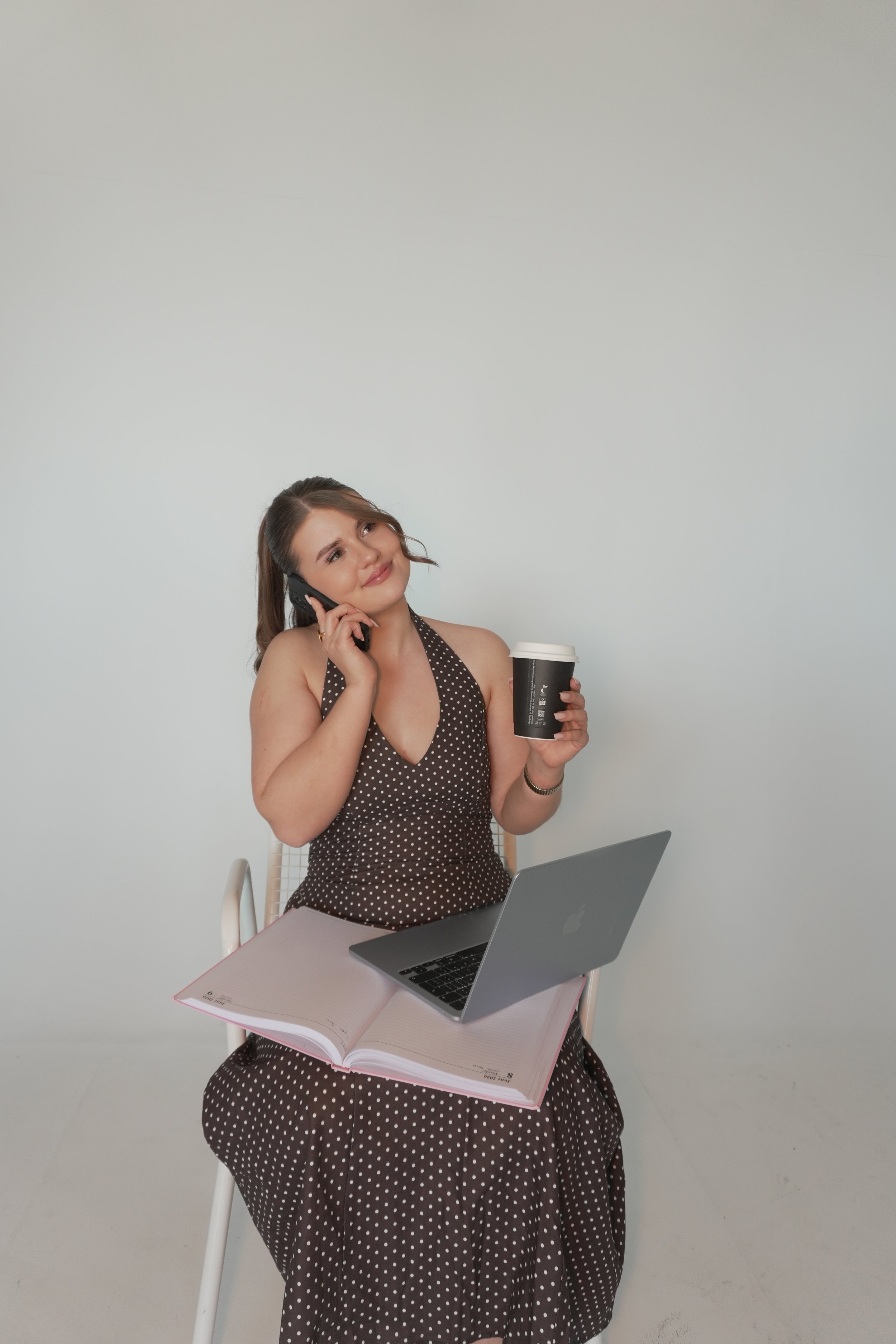 A woman in a polka dot dress is sitting at a table, holding a cup of coffee in one hand and a cell phone to her ear with the other. She has a laptop and an open planner in front of her. The background is plain and white.