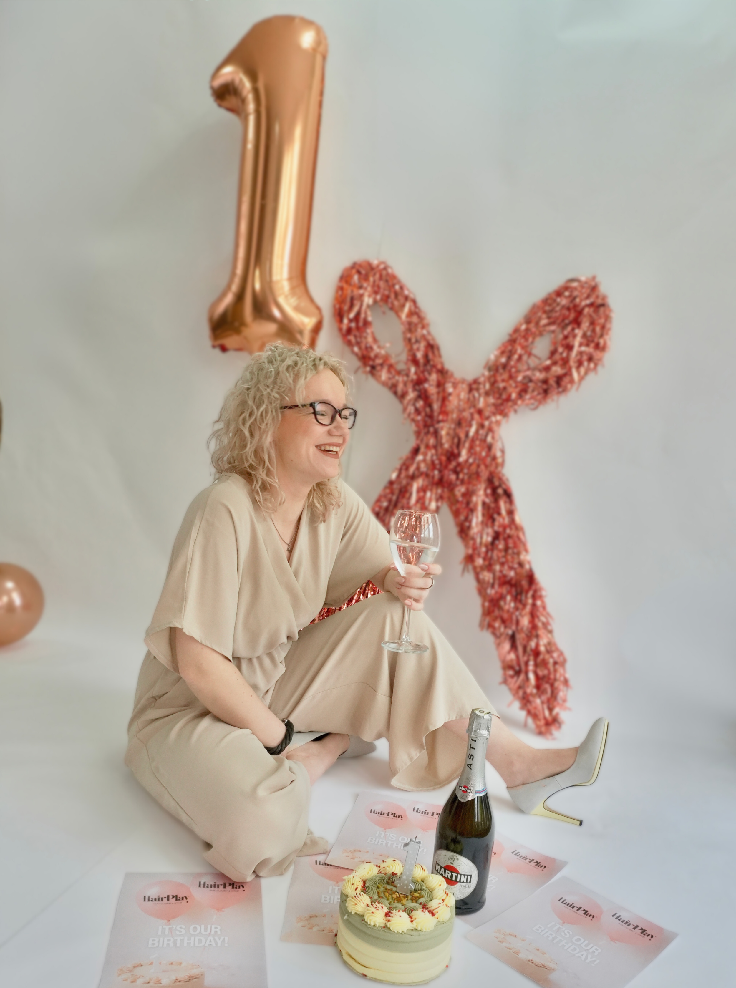 A woman with curly blonde hair and glasses sitting on the floor holding a glass of champagne, with birthday decorations behind her including a gold balloon shaped as the number 1 and a sequined red balloon shaped as the number 1, a cake with a candle