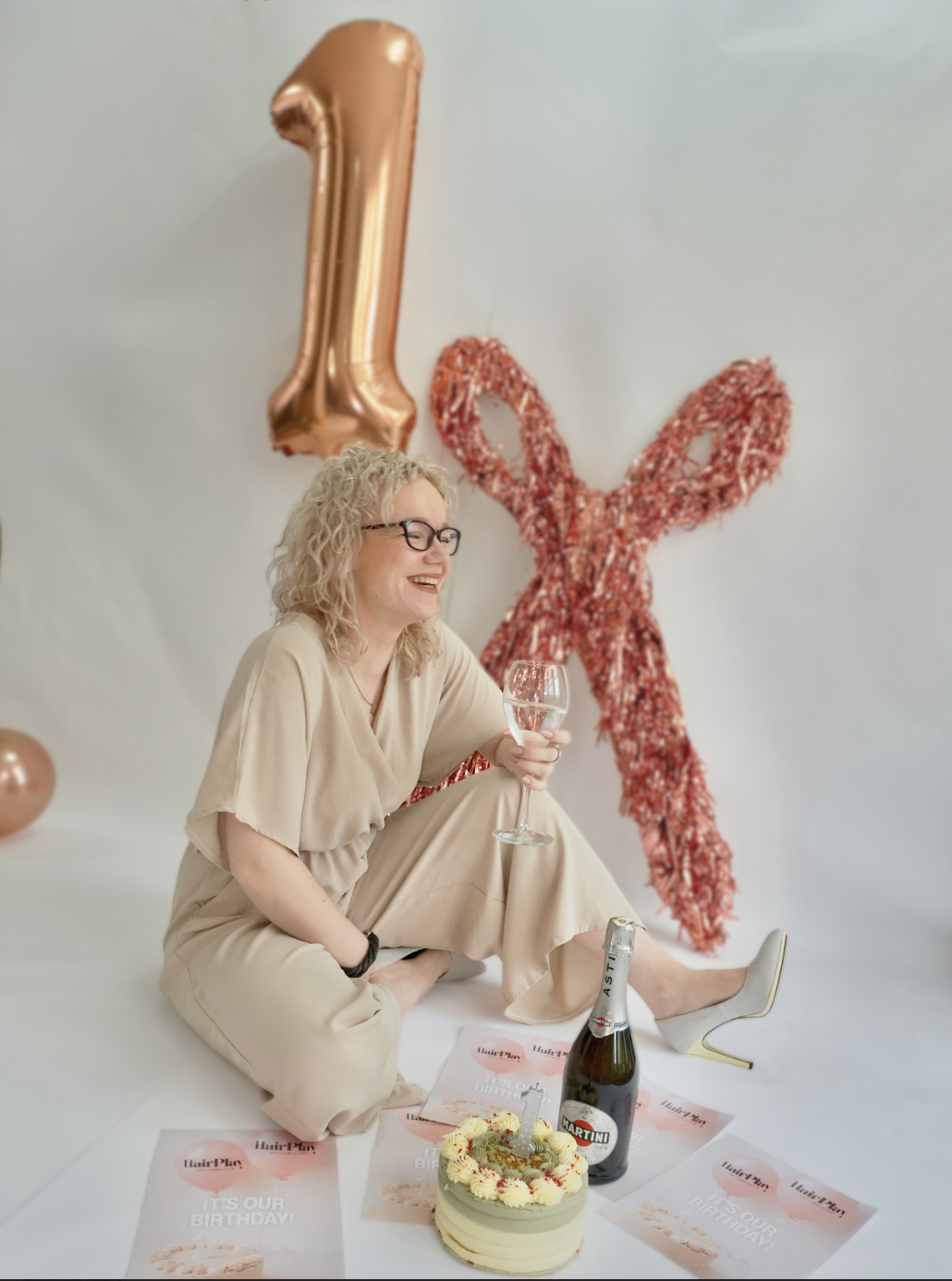 Woman celebrating her first birthday, sitting on the floor with a glass of water, surrounded by birthday decorations, a cake, and a bottle of Martin wine.