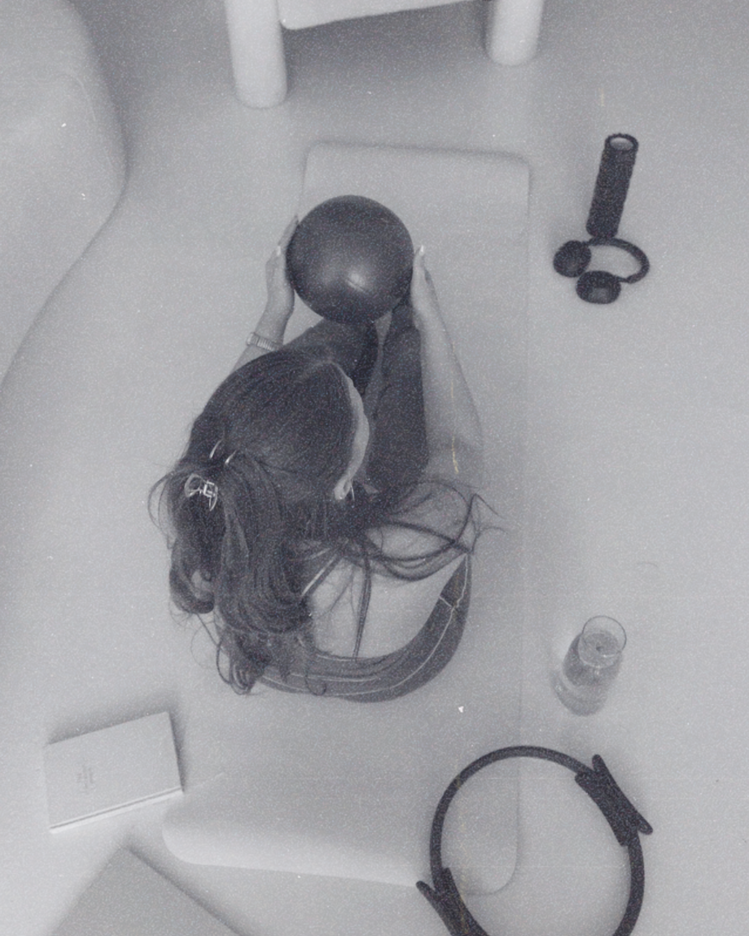 A person holding a black medicine ball standing on a white tiled floor in a room with a white chair, a pair of black headphones, and a black exercise resistance band nearby.