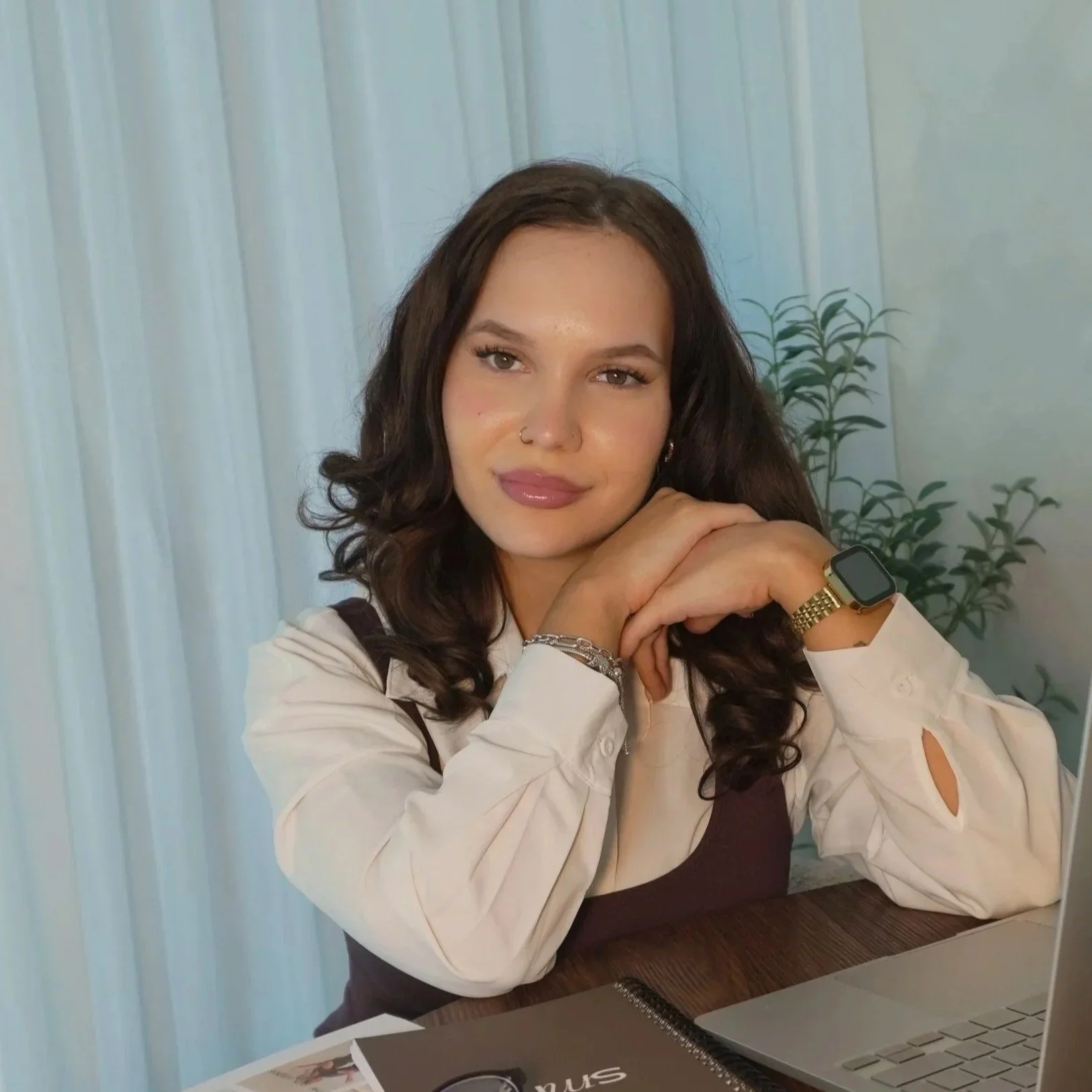 A young woman with long curly dark hair, wearing a white long sleeve shirt with cutouts on the sleeves, jewelry, a smartwatch, and a nose ring, sitting at a table with a laptop, a notebook, and a printed paper in front of her. She is resting her chin on her folded hands and looking into the camera with a slight smile.