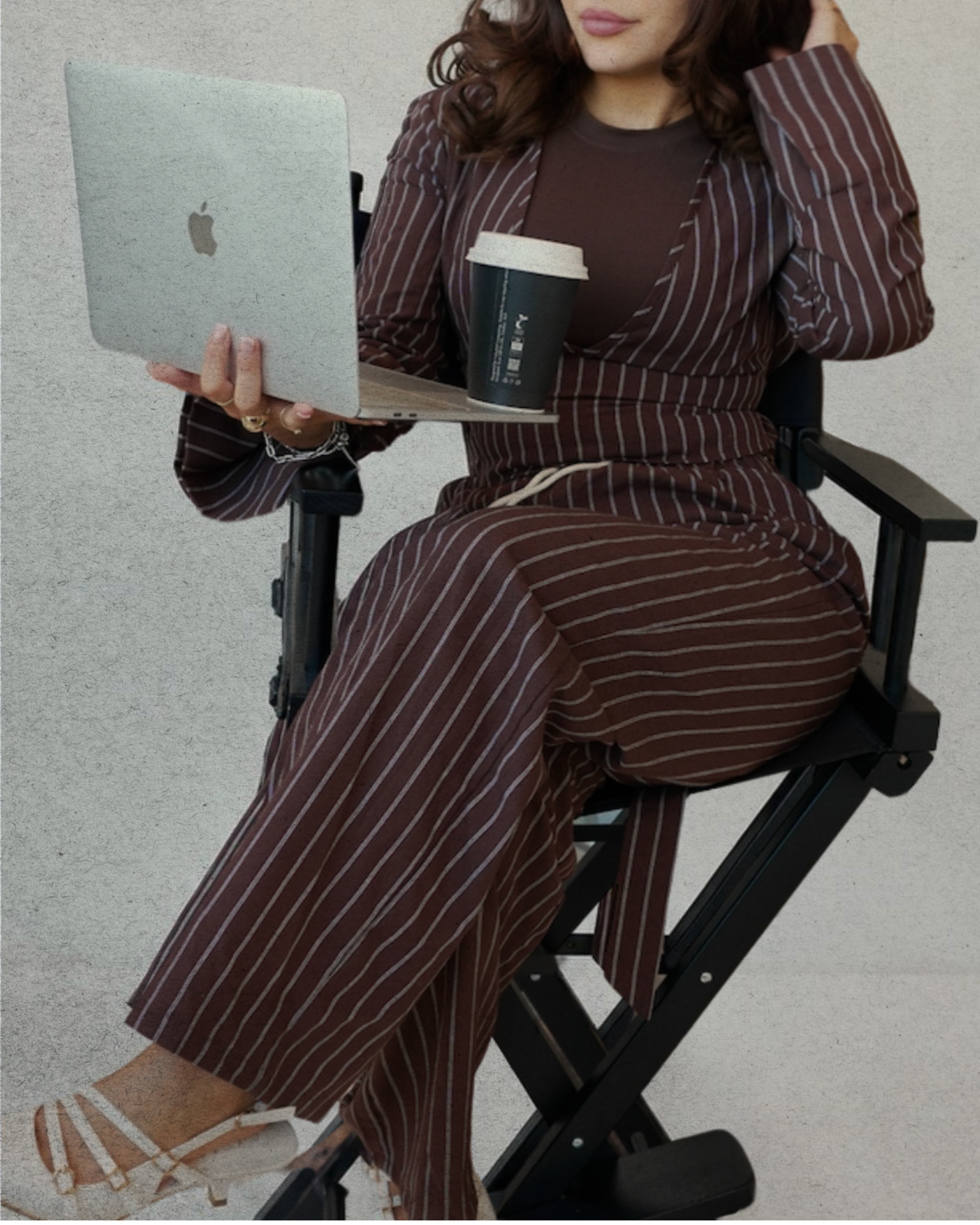 A woman sitting on a black folding chair outdoors, holding a silver MacBook and a black take-out coffee cup, wearing a brown striped dress and white sandals, with a gray wall in the background.