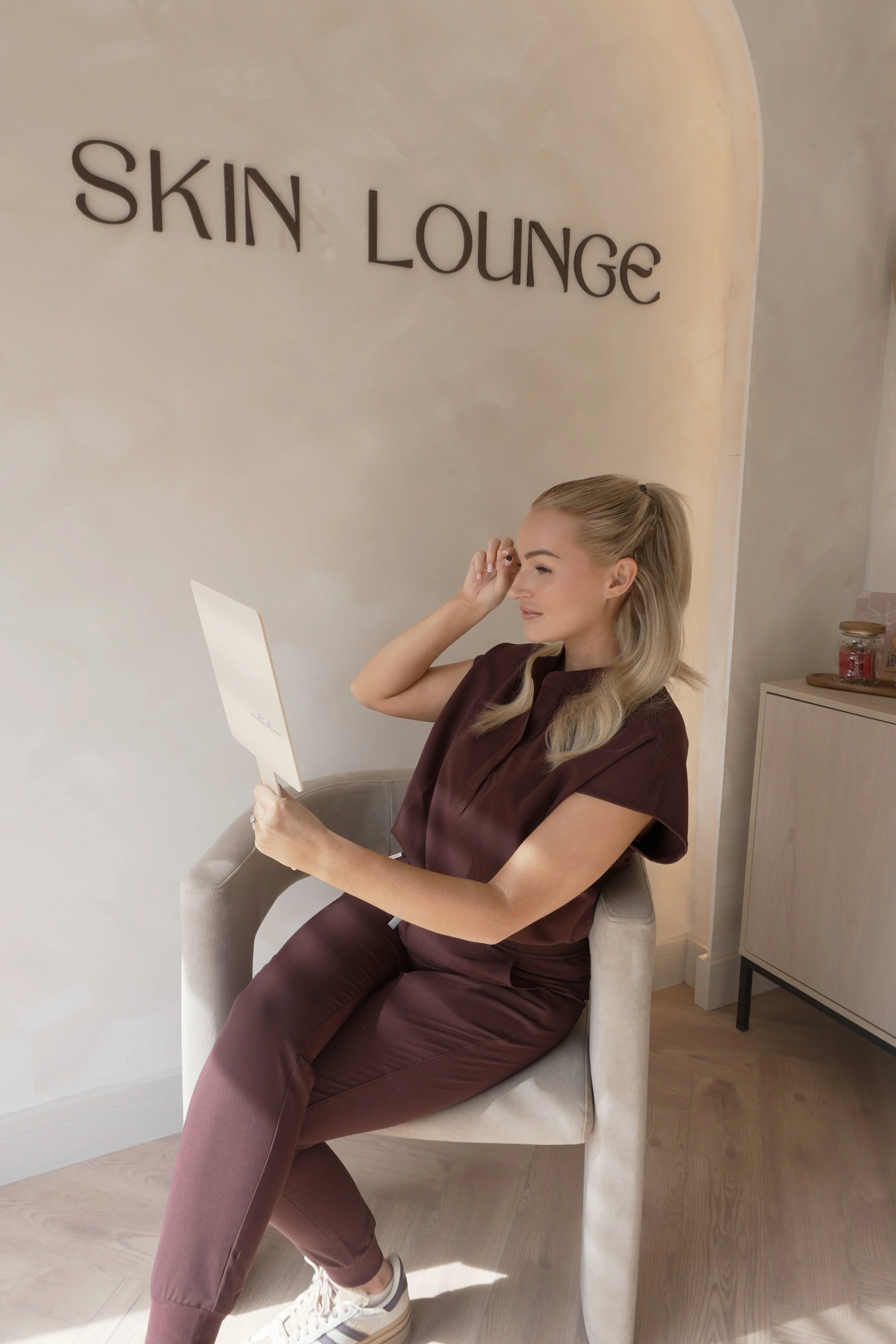 A woman sitting in a beige chair, wearing a matching brown skincare professional outfit, holding a paper or card, and sitting inside a skincare lounge with a sign that reads 'SKIN LOUNGE' on the wall behind her.